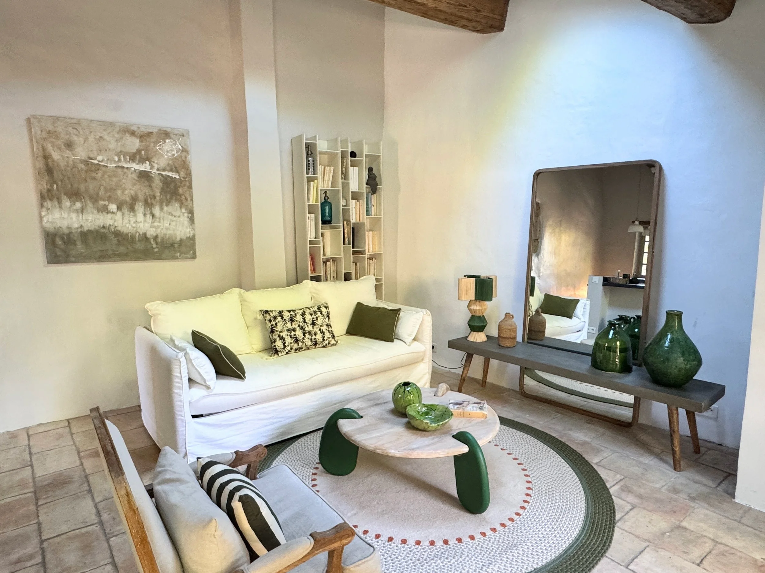 Un salon avec un canapé blanc, des coussins décoratifs, une étagère blanche, un grand miroir, des vases verts, une table en bois, un tapis rond avec une bordure rouge, et un mur décoré d'une peinture abstraite.