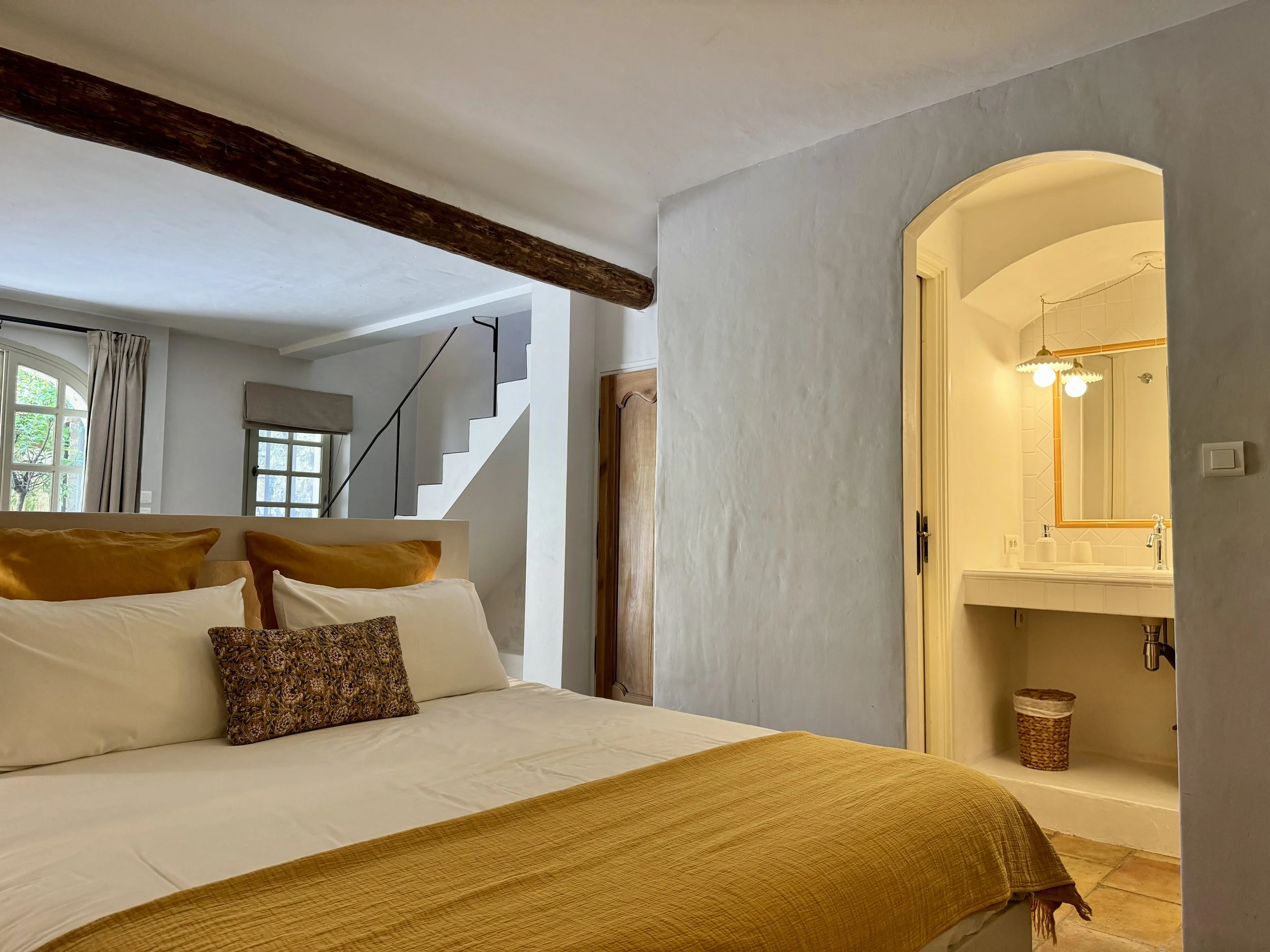 Une chambre avec un lit, des coussins, une fenêtre avec rideaux, une porte en bois, un escalier blanc, un plafond avec une poutre en bois, et une salle de bain avec un miroir, un lavabo, et un éclairage