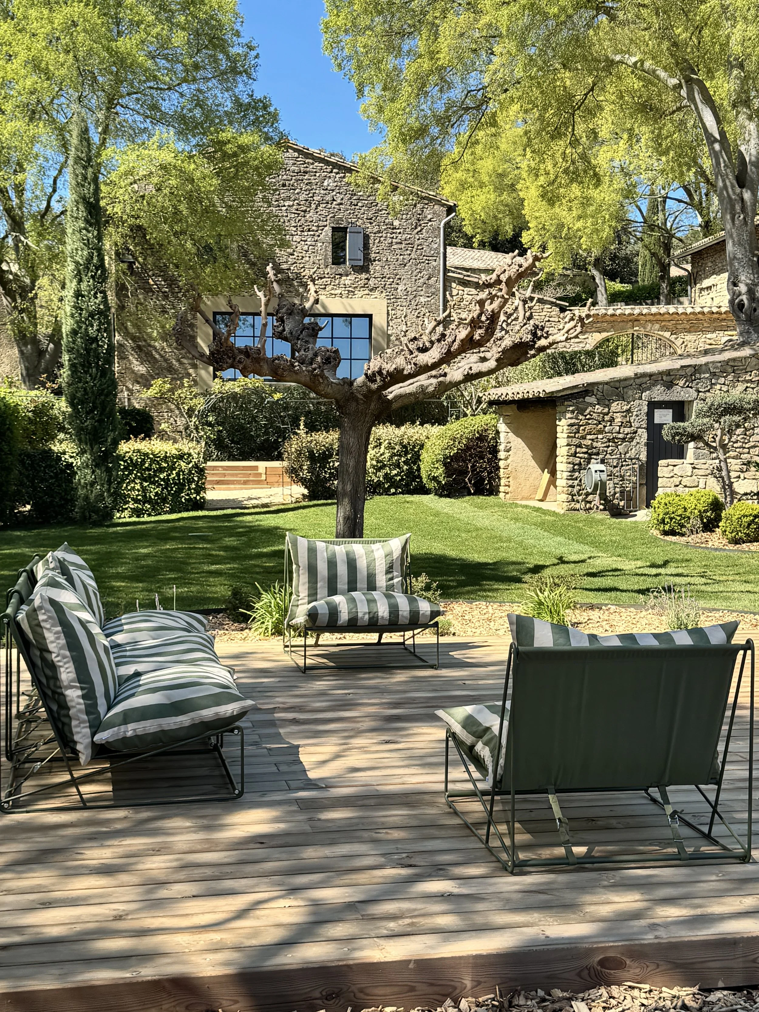 Terrasse en bois avec des meubles en métal et des coussins rayés, jardin avec pelouse, arbres et vieille maison en pierre en arrière-plan, ciel bleu ensoleillé.