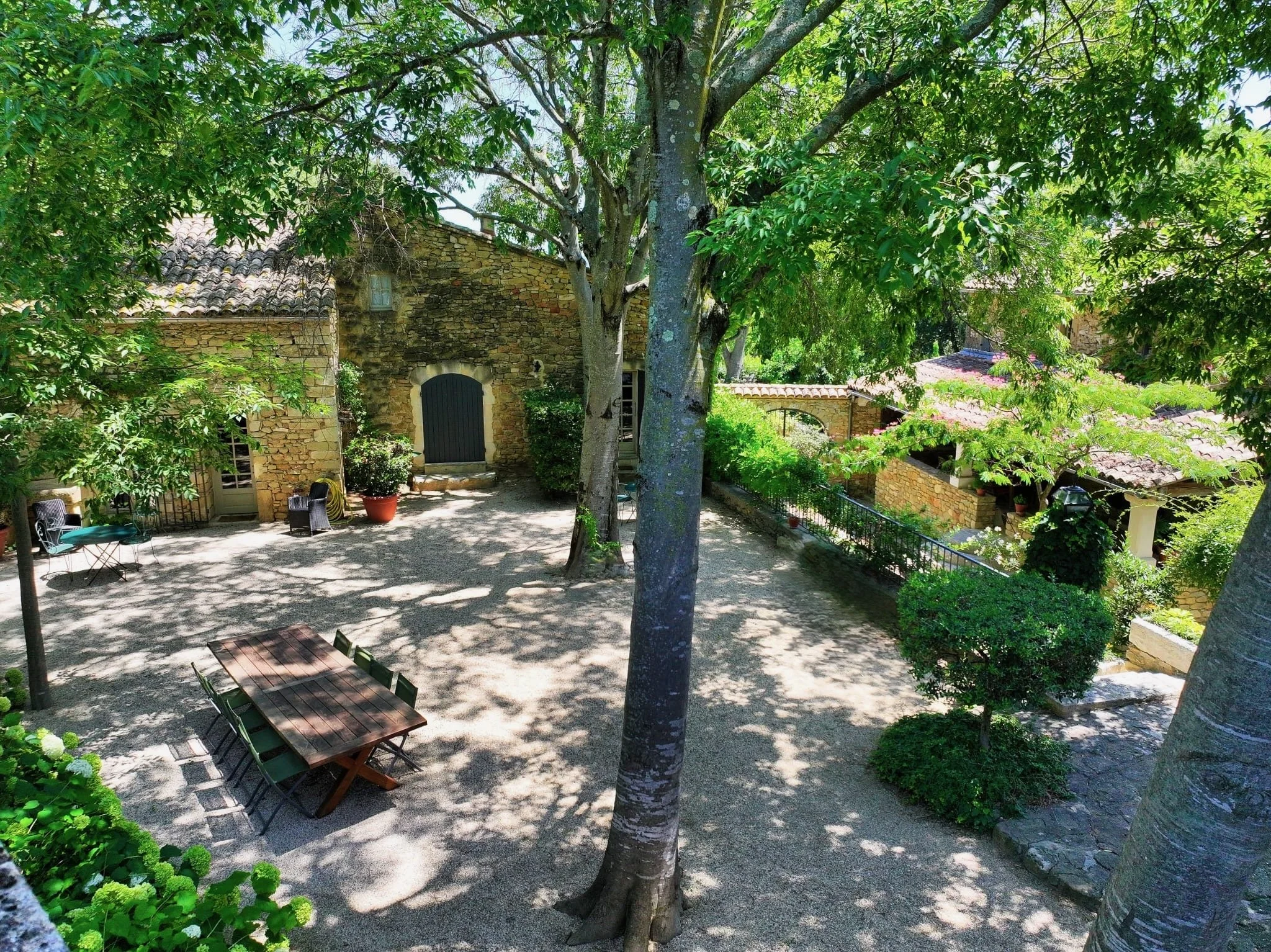 une terrasse du sud qui ressemble à une place de village avec des batiments anciens, de grands arbres, une table de repas