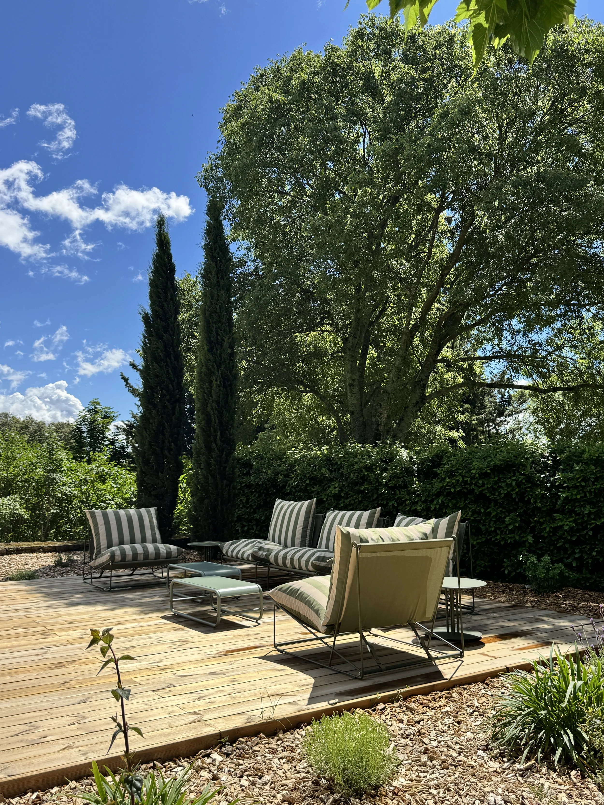 Une terrasse en bois avec des meubles de jardin à rayures vertes et blanches, entourée de végétation dense, avec de grands arbres et un ciel bleu en arrière-plan.