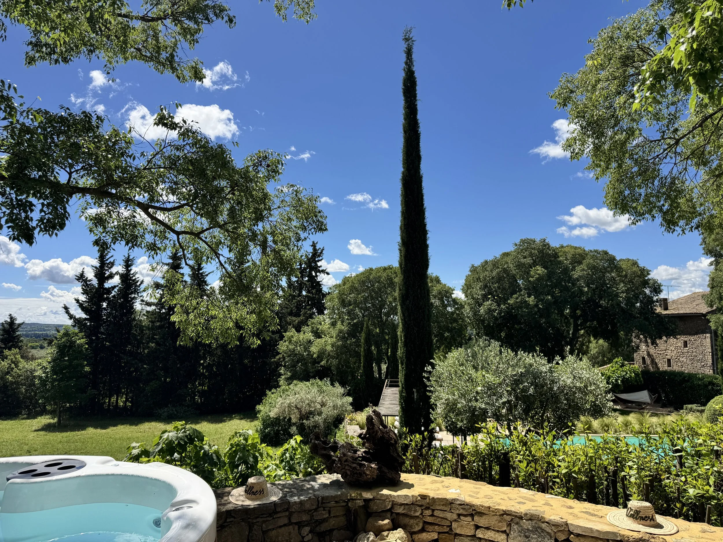 Jardin avec piscine, arbres verts, ciel bleu avec quelques nuages, bâtiment ancien en pierre. 