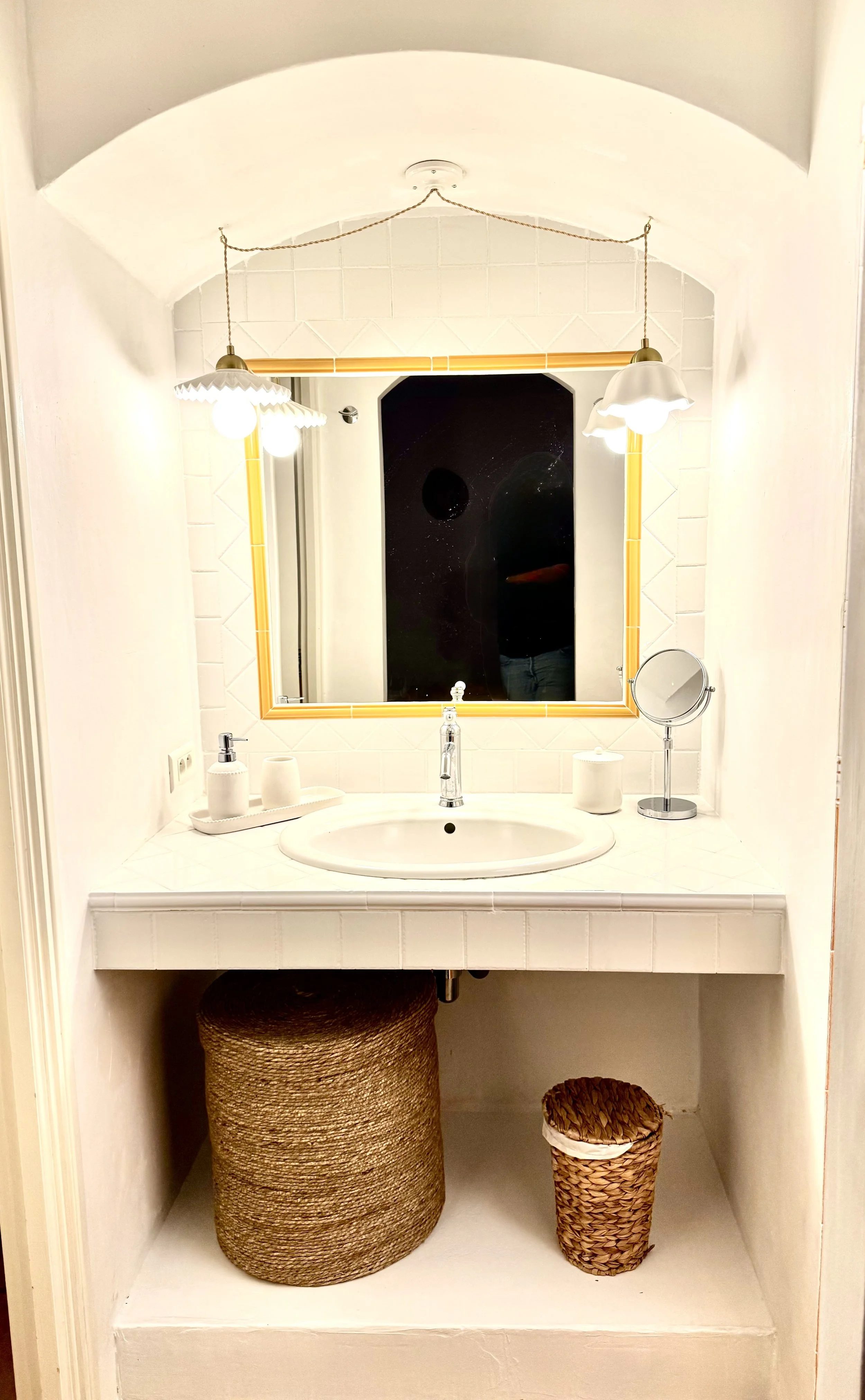 Lavabo avec miroir, éclairage, et accessoires de salle de bain dans un espace blanc, avec paniers en dessous.