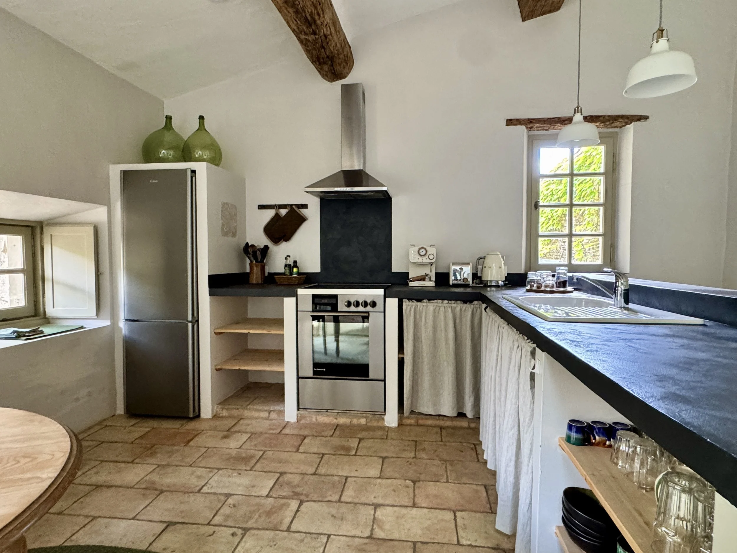 Cuisine de style rustique avec une fenêtre laissant entrer la lumière, comprenant un réfrigérateur, une cuisinière à gaz, une hotte en acier inoxydable, des étagères avec des ustensiles et des verreries, et un plan de travail blanc avec un rideau, ca