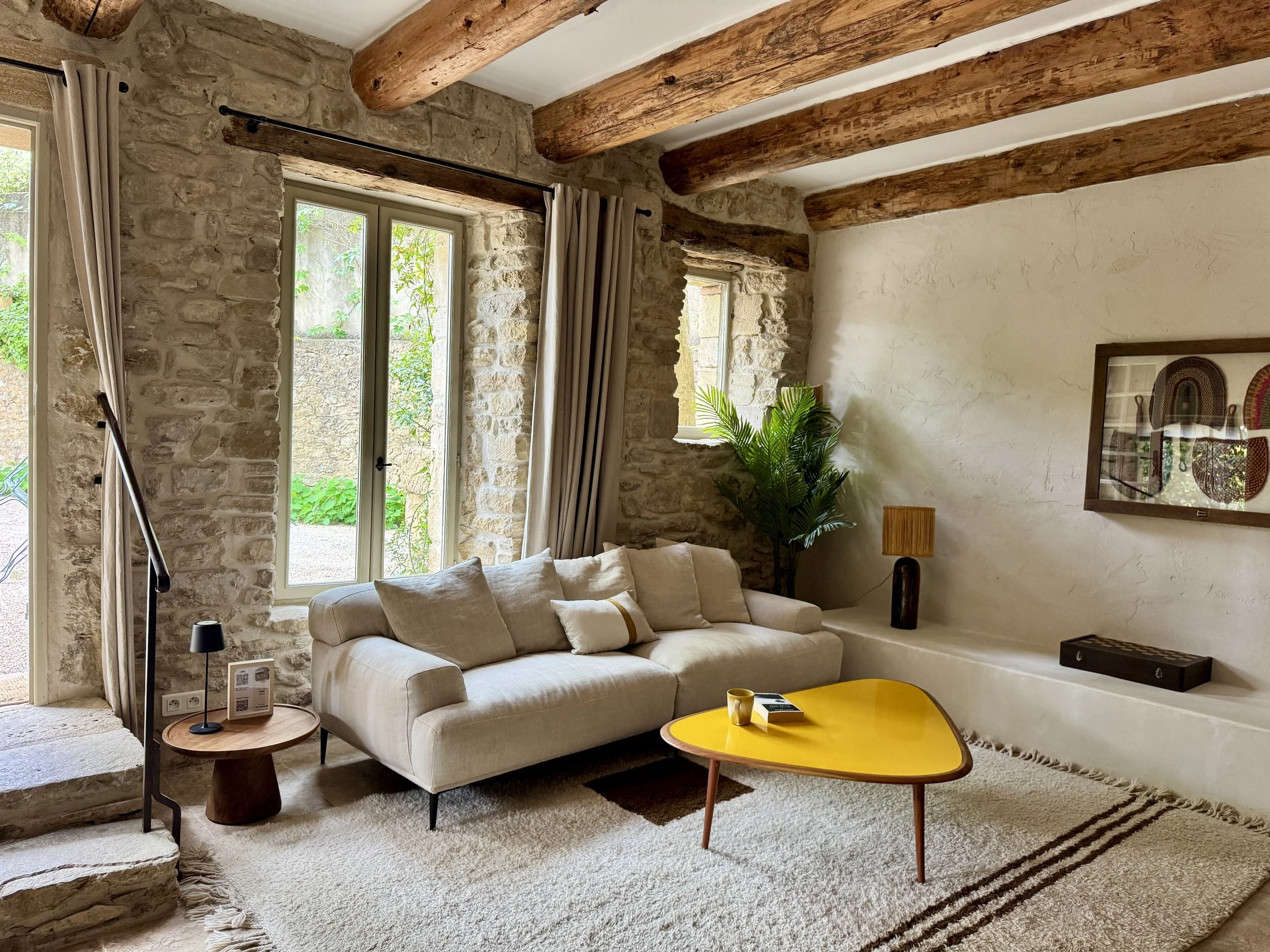 Salon avec canapé beige, murs en pierre, poutres en bois au plafond, grande fenêtre avec rideaux, tableau mural, plante verte, tapis, table basse jaune, lampe de lecture