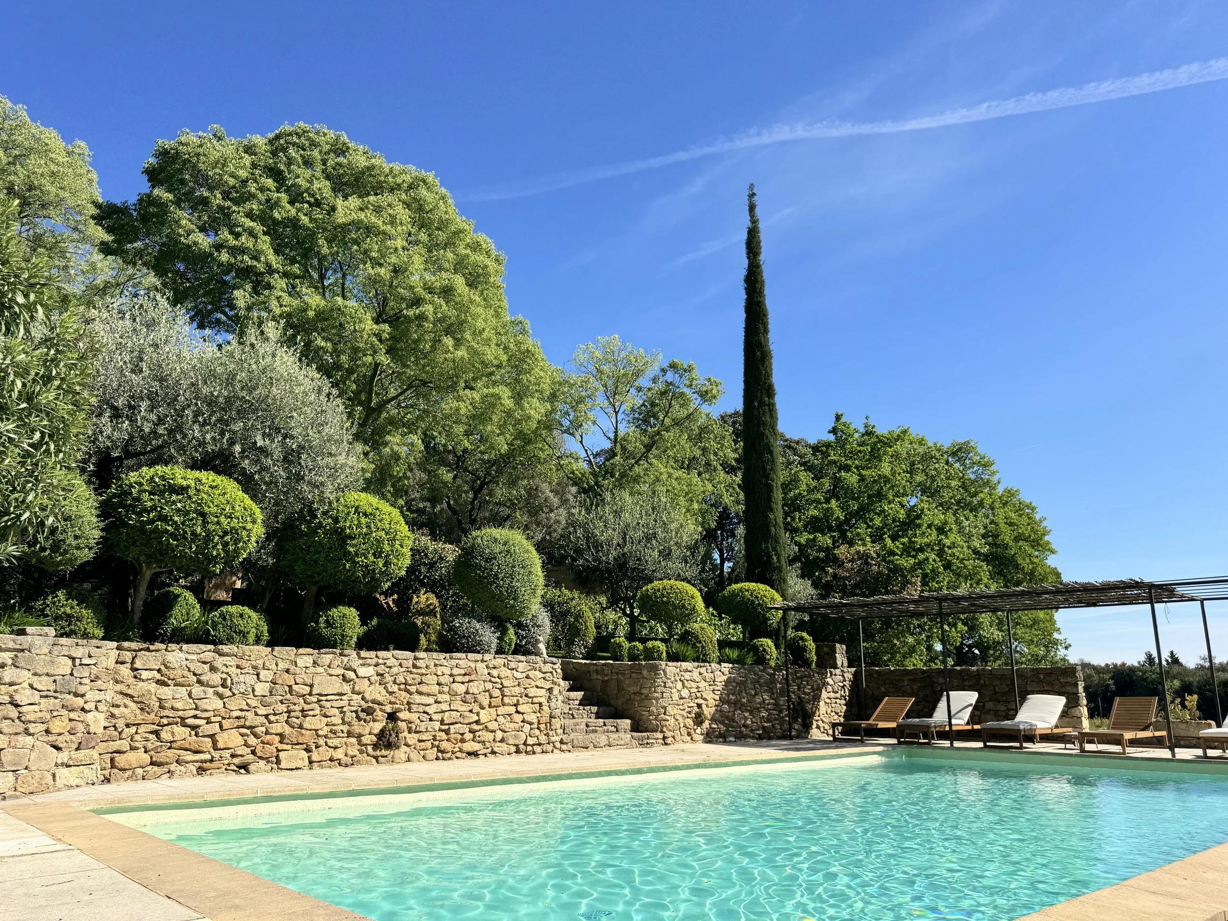 Piscine extérieure avec chaises longues, mur en pierre, arbres verts et ciel bleu en arrière-plan.