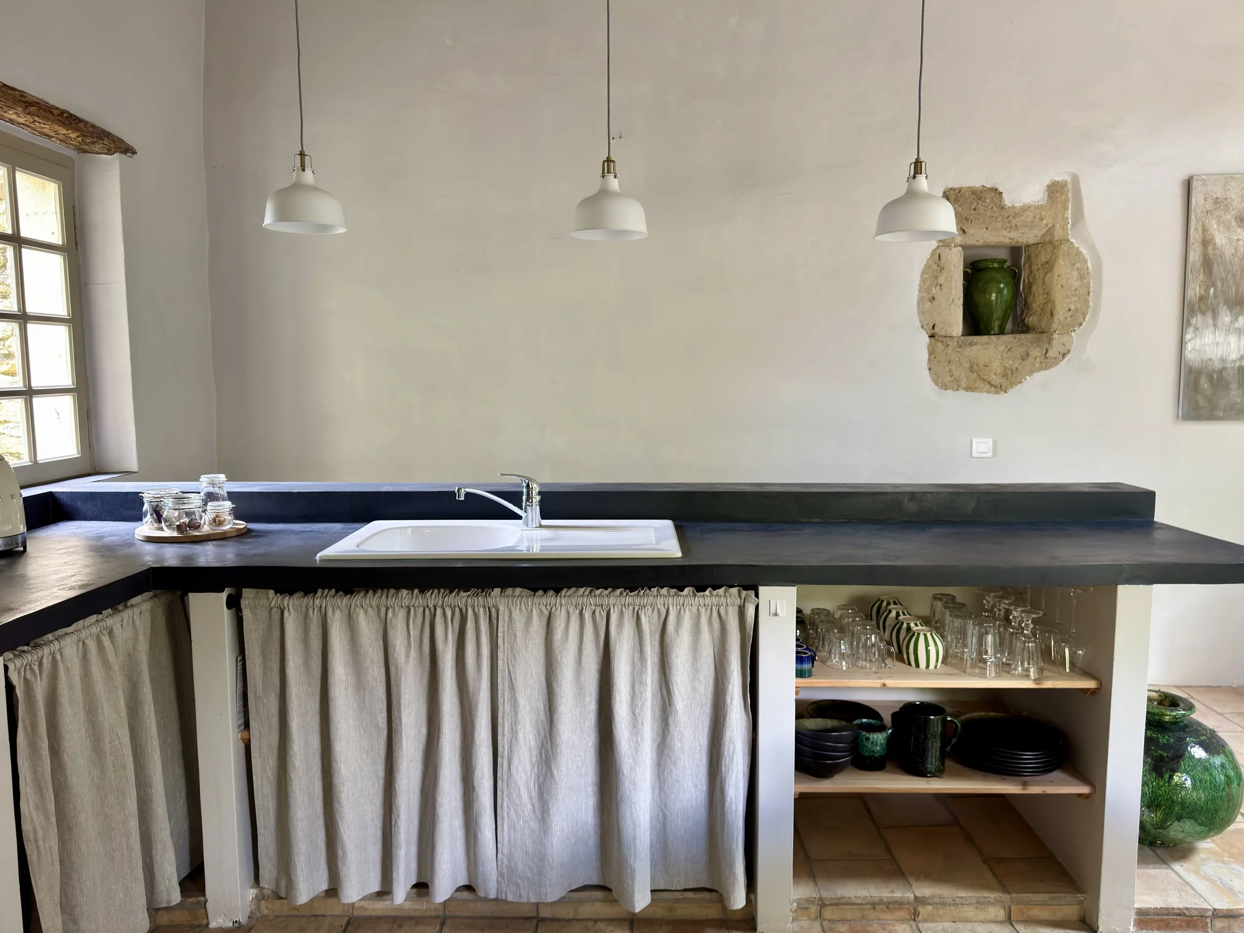 Une cuisine avec un comptoir en pierre noire, un évier blanc, trois lampes suspendues blanches au plafond, une fenêtre laissant entrer la lumière, et un mur en pierre avec un nichoir contenant une grande poterie verte. En dessous du comptoir, un ride