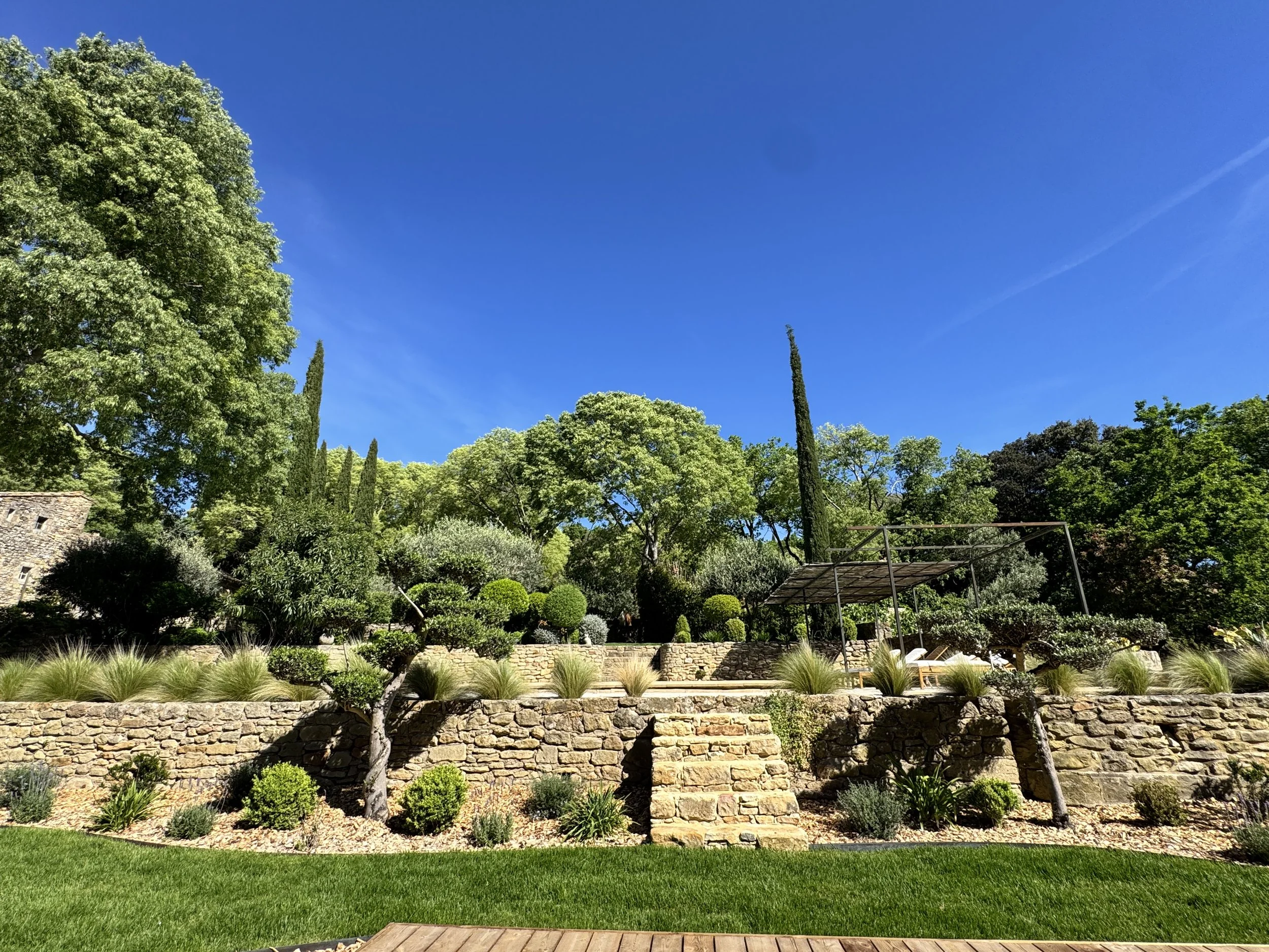 Jardin bien entretenu avec pelouse verte, buissons, arbres, un mur en pierre, un parterre en contrebas, une structure en métal et un ciel bleu clair.