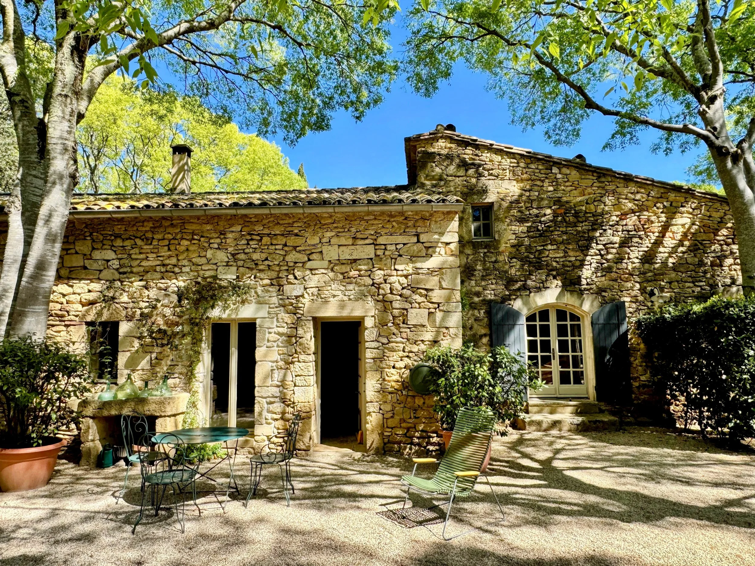 Une maison en pierre avec fenêtres à volets bleus, entourée d'arbres verts sous un ciel bleu clair.
