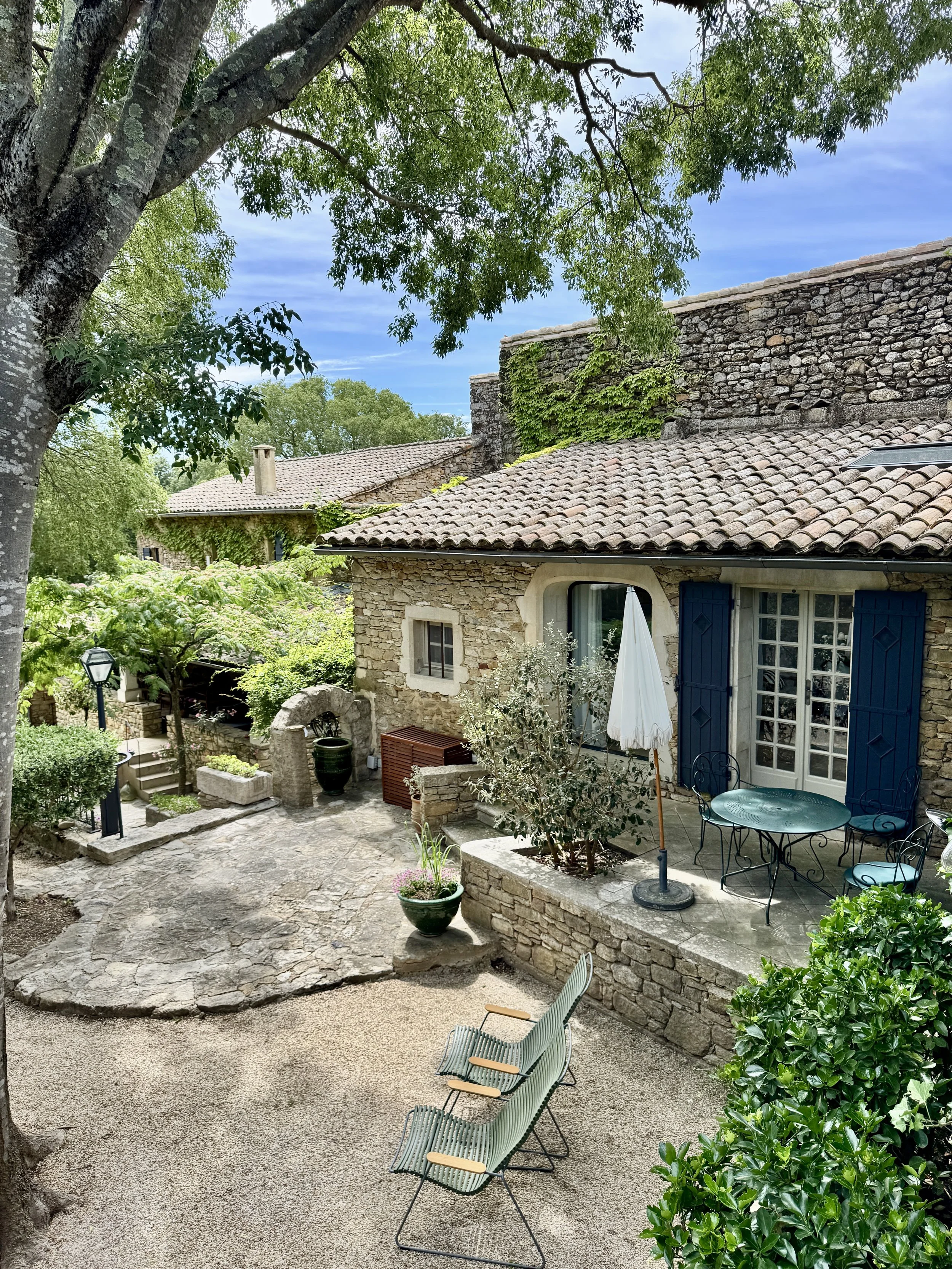 Jardin avec des chaises en métal vertes, une table ronde en métal, une grande maison en pierre avec des volets bleus, des plantes en pot, une grande arbre avec feuillage dense, un mur en pierre avec de la végétation et des escaliers en pierre menant 