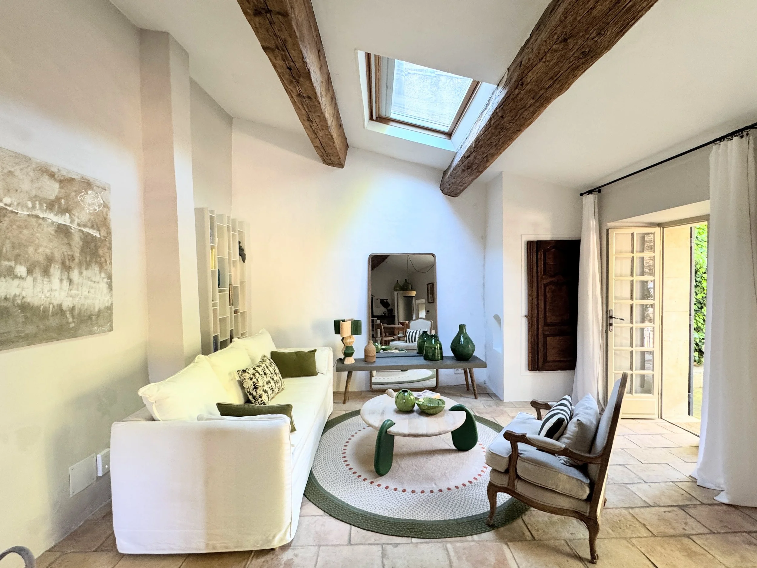 Intérieur d'un salon avec un plafond à poutres en bois, une fenêtre de toit, un canapé blanc, une table basse ronde, et des fauteuils en bois avec coussins rayés, décoré avec des vases en verre vert.