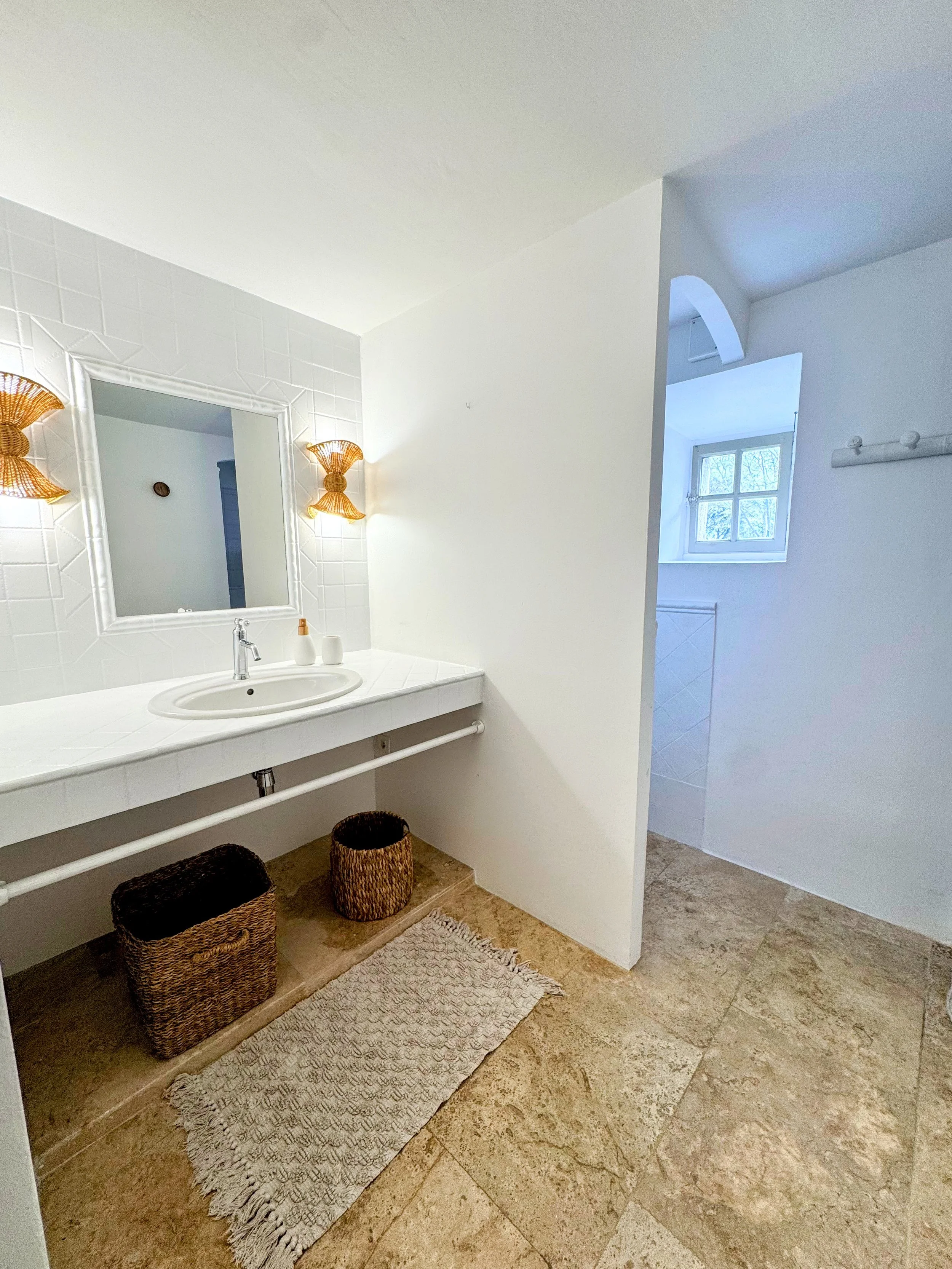 Vue intérieure d'une salle de bain avec un lavabo, un miroir, deux appliques murales en osier, un tapis en laine, et une fenêtre avec vue sur l'extérieur.
