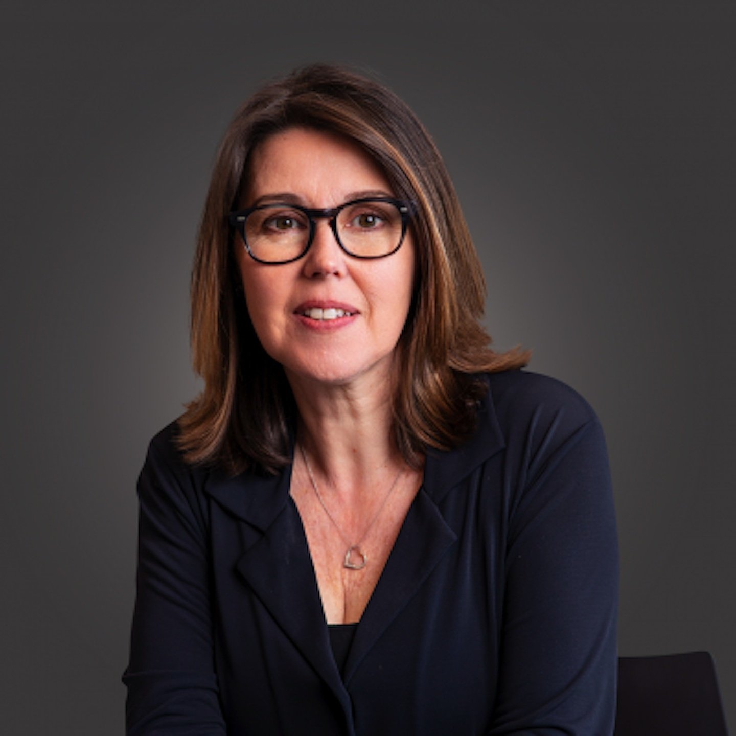 Headshot of a woman with shoulder-length brown hair, wearing black glasses and a black top, against a dark gray background.