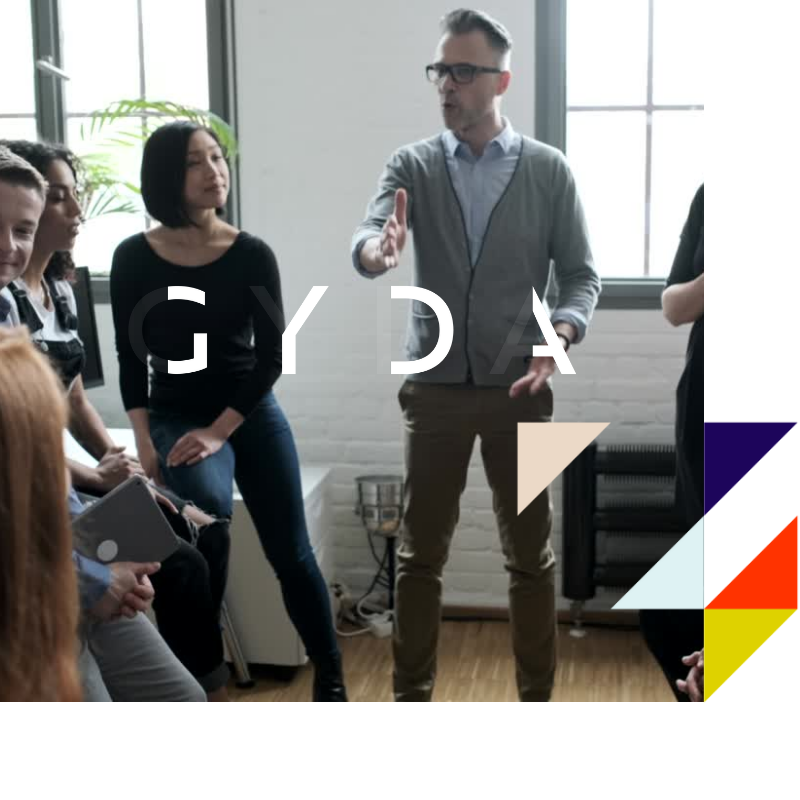 A man with glasses and gray sweater giving a presentation or speech to a diverse group of people in a bright office setting.