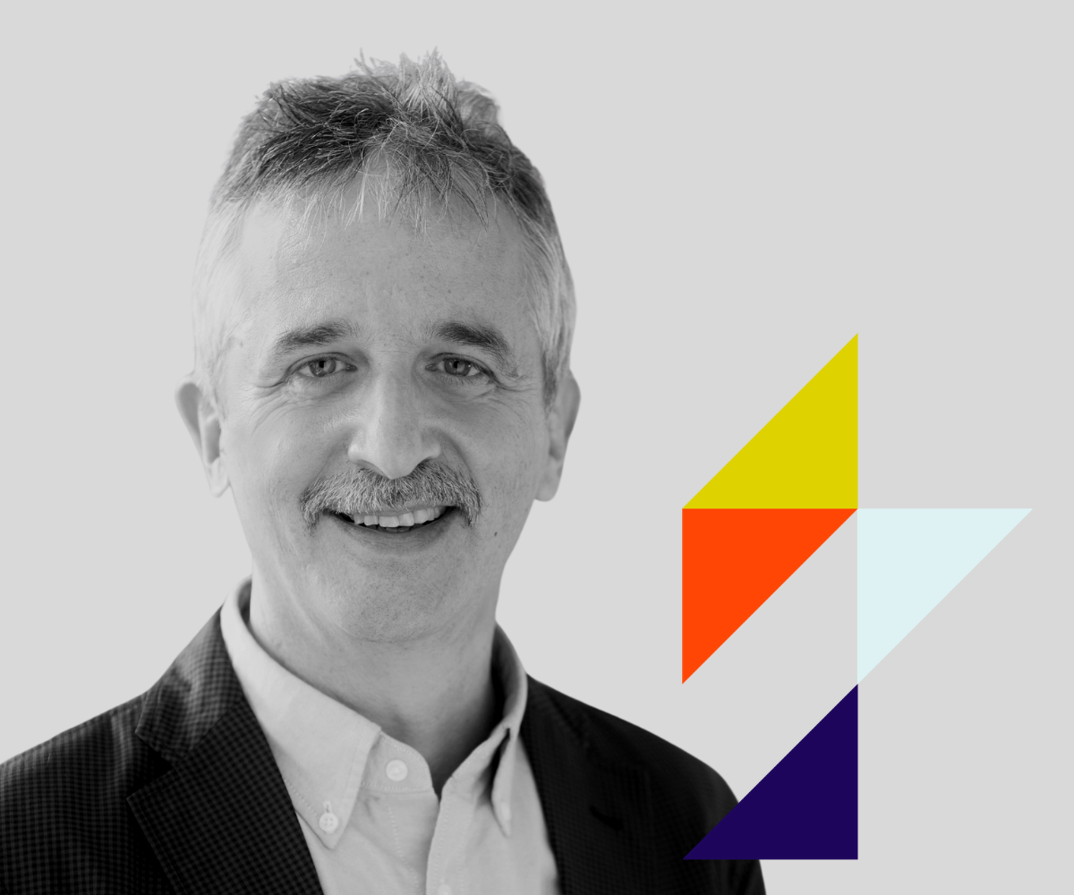 A black and white portrait of a smiling man with light-colored hair and a mustache, wearing a checkered blazer and a white shirt, with geometric colored shapes on the right side of the image.