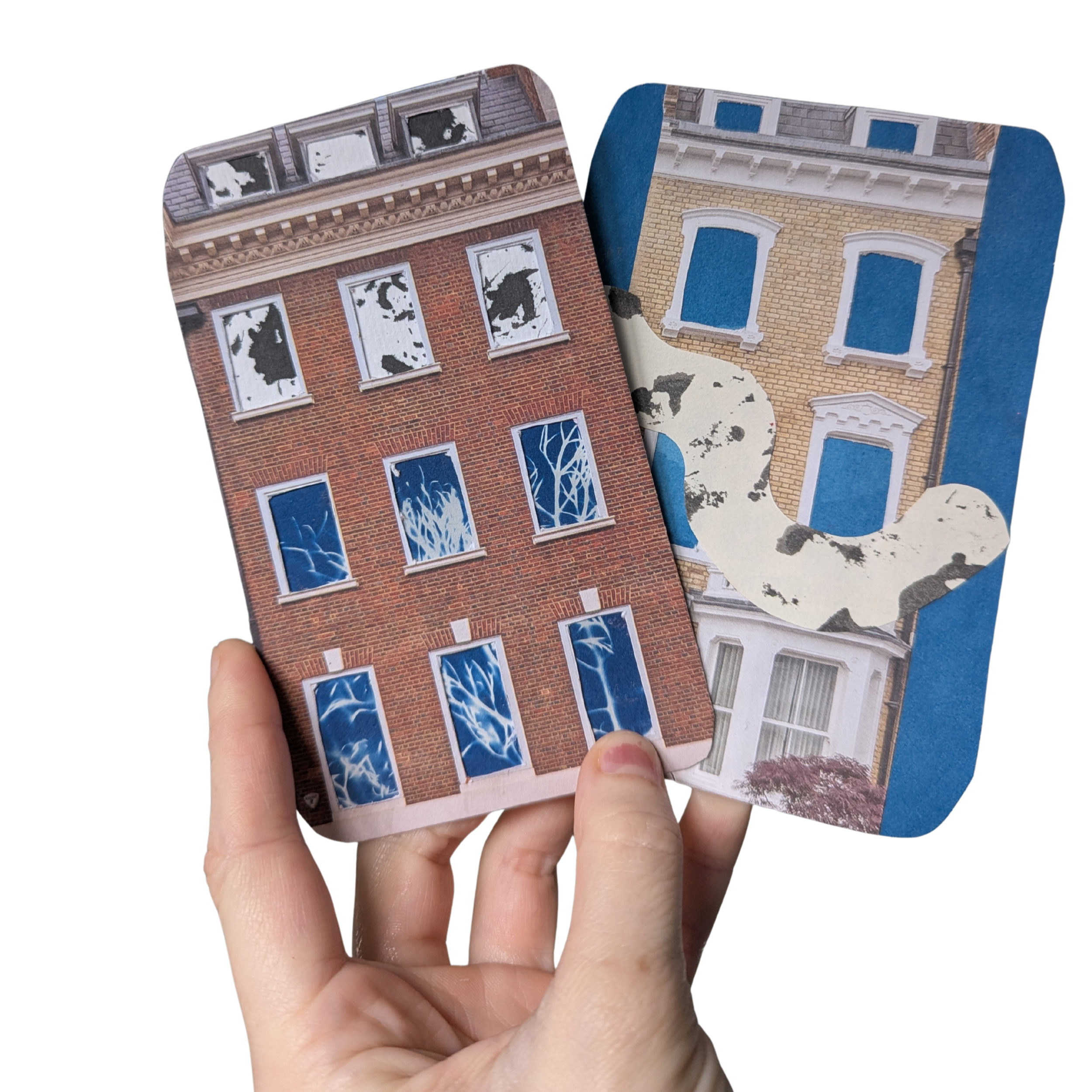 My hand is holding 2 artist trading cards. Both cards have cut out house fronts on top of cyanotype and mark-making prints which you can see through the cut out windows.