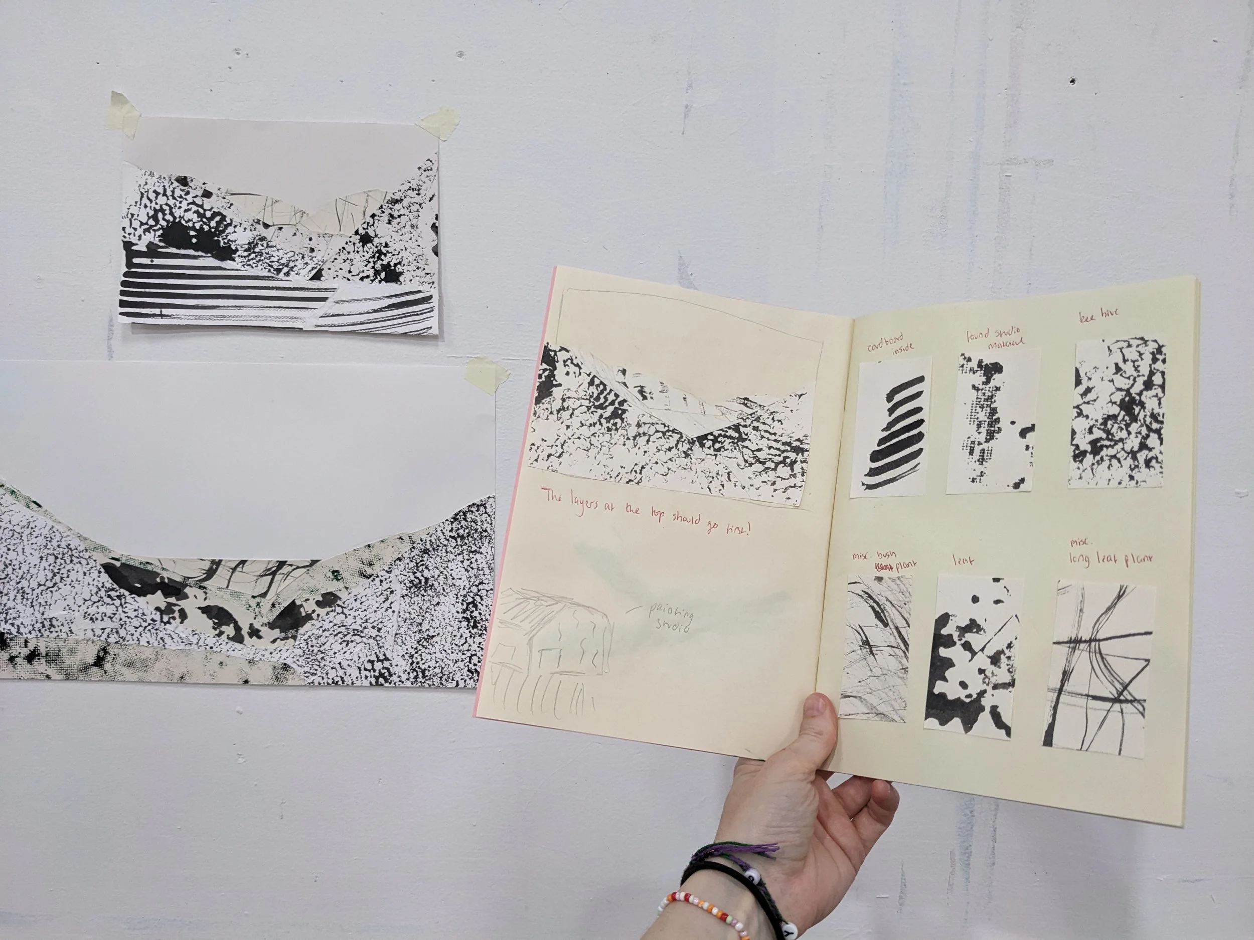 holding up a sketchbook that shows the documentation of the different marks and what they are. Some mark-making landscapes are shown in the background.