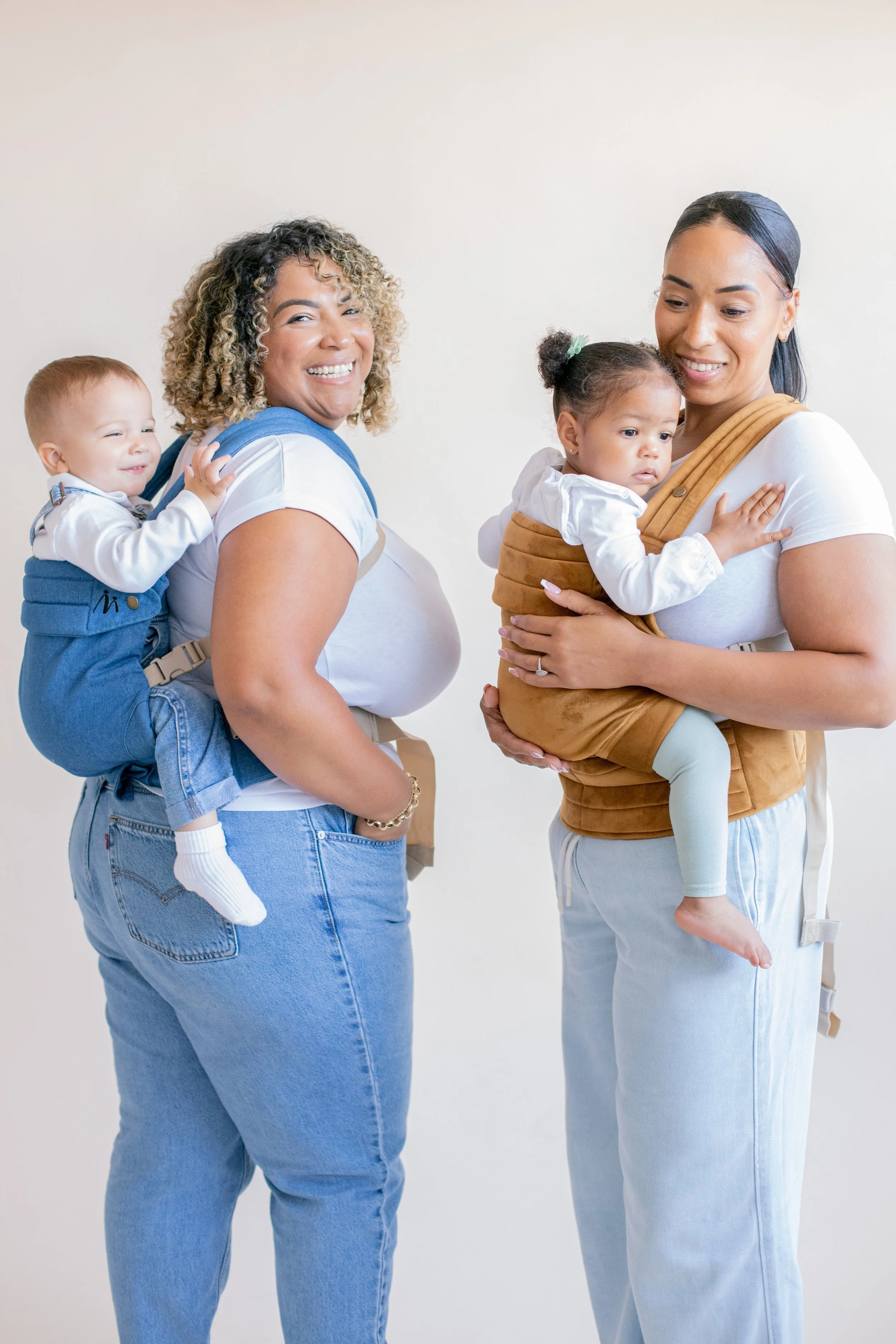 Two women holding young children in baby carriers, standing side by side against a plain white wall. The woman on the left has curly blonde hair and is smiling, while the woman on the right has straight dark hair and is looking down at the child she is holding.