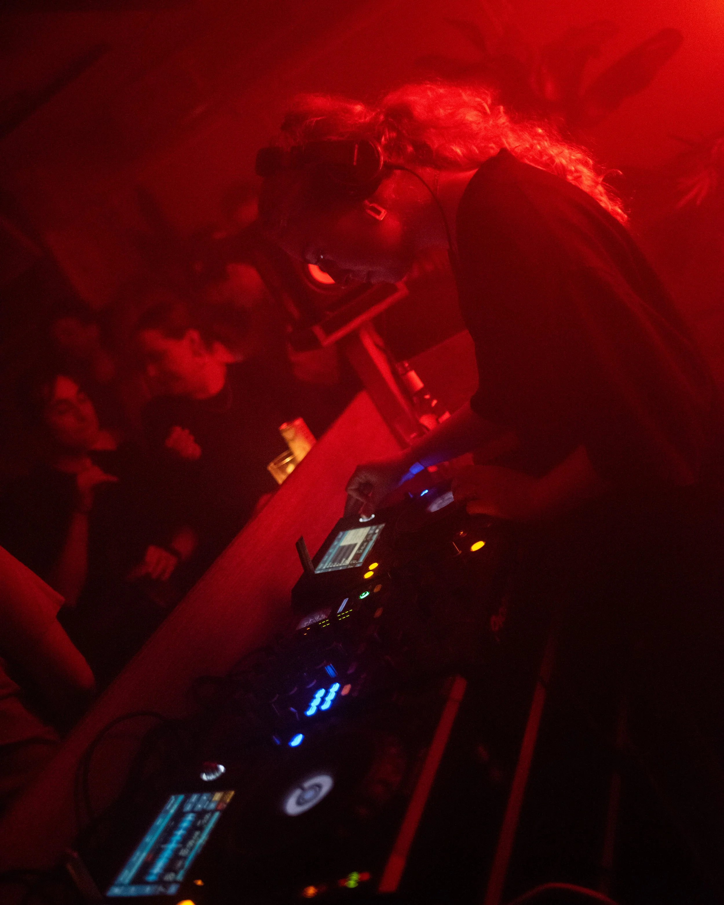DJ performing in a nightclub with red lighting, surrounded by sound equipment, and an audience in the background.
