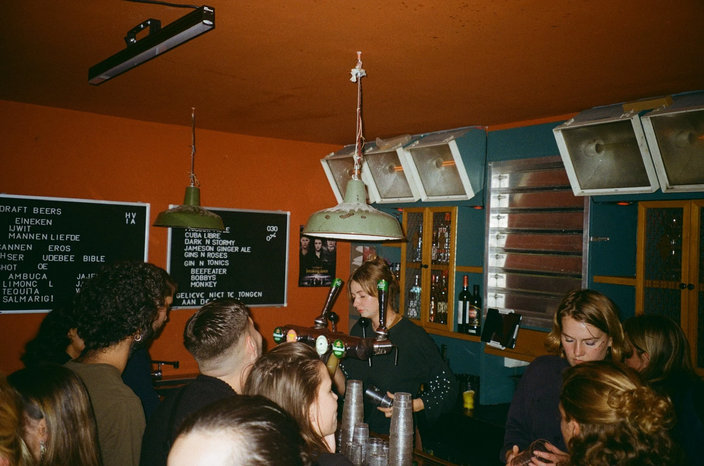 Crowded bar with bartender serving drinks, menu on wall, and people socializing