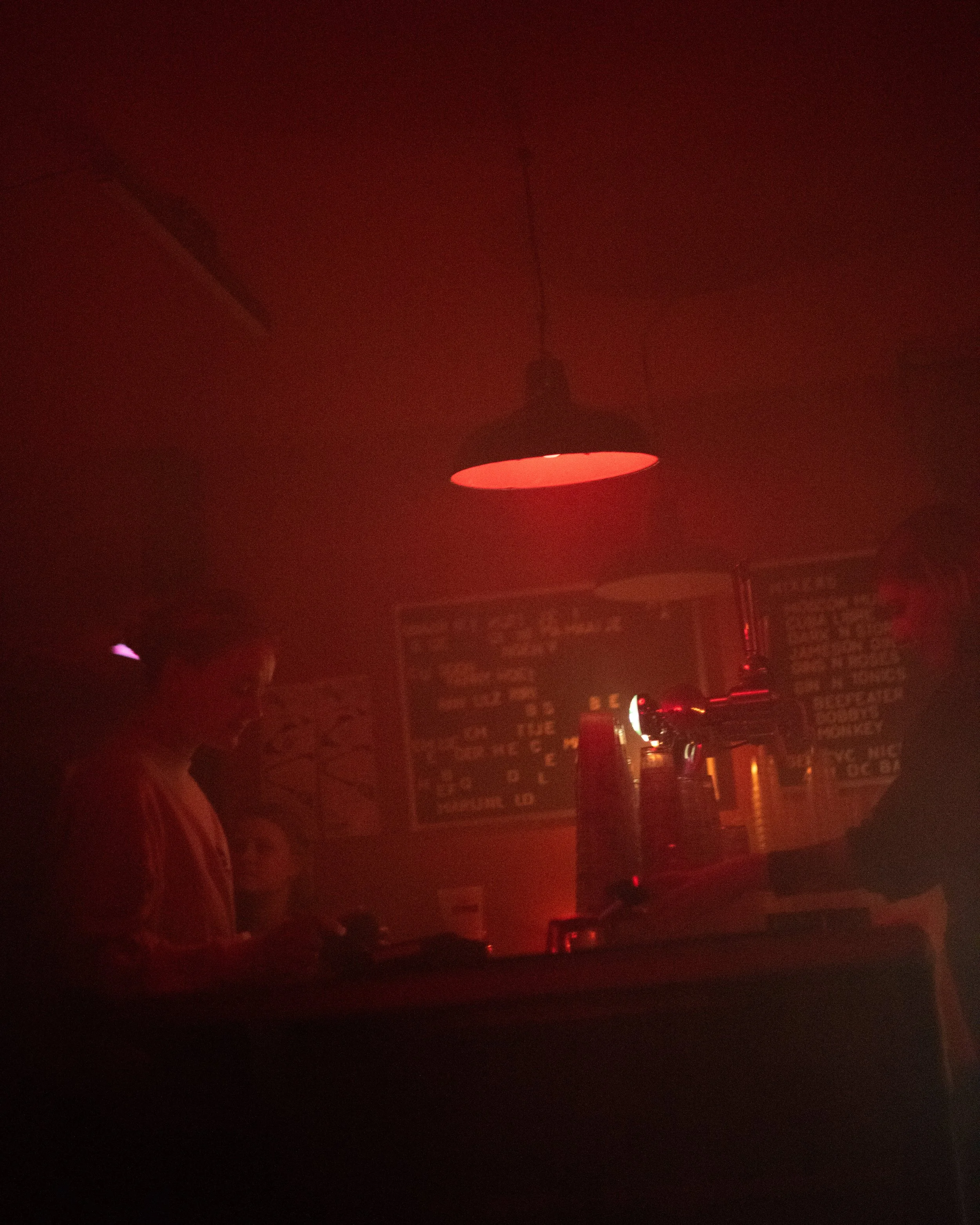 Dimly lit bar interior with people, red lighting, and a chalkboard menu in the background.