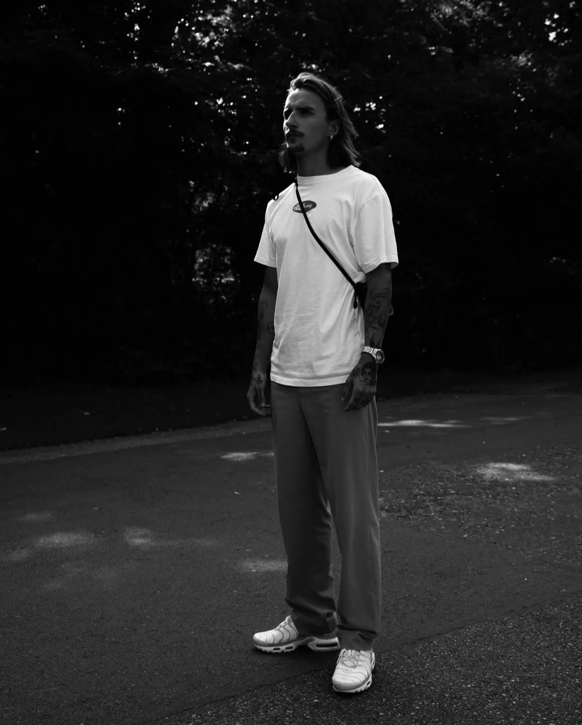 Black and white photo of a man standing on a road, wearing a white t-shirt, pants, and sneakers, with trees in the background.