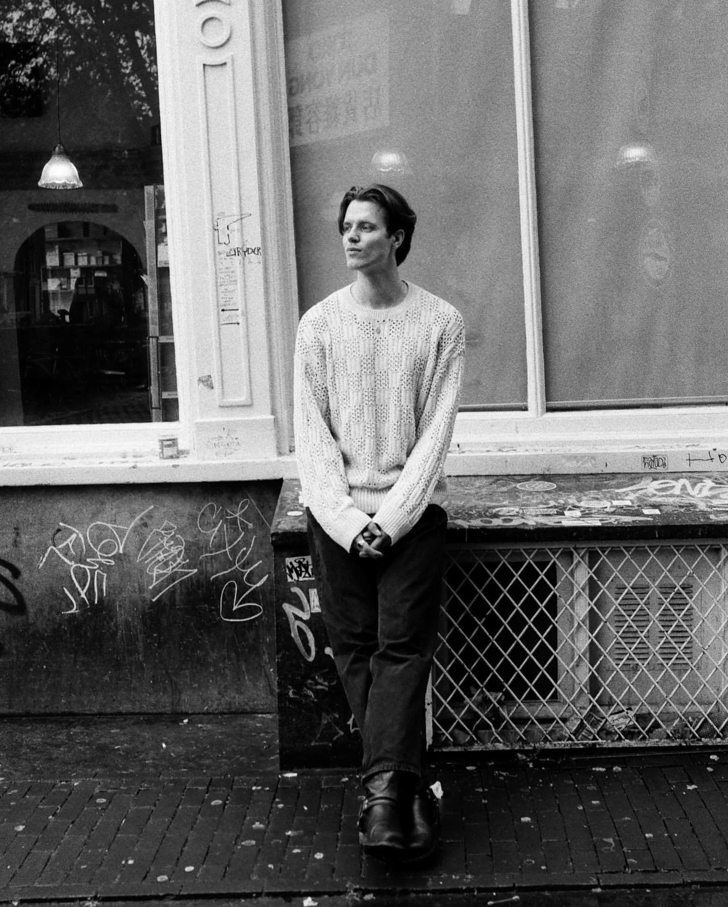 Black and white photo of a person in a knitted sweater standing against a building exterior, with graffiti on the wall and window nearby.