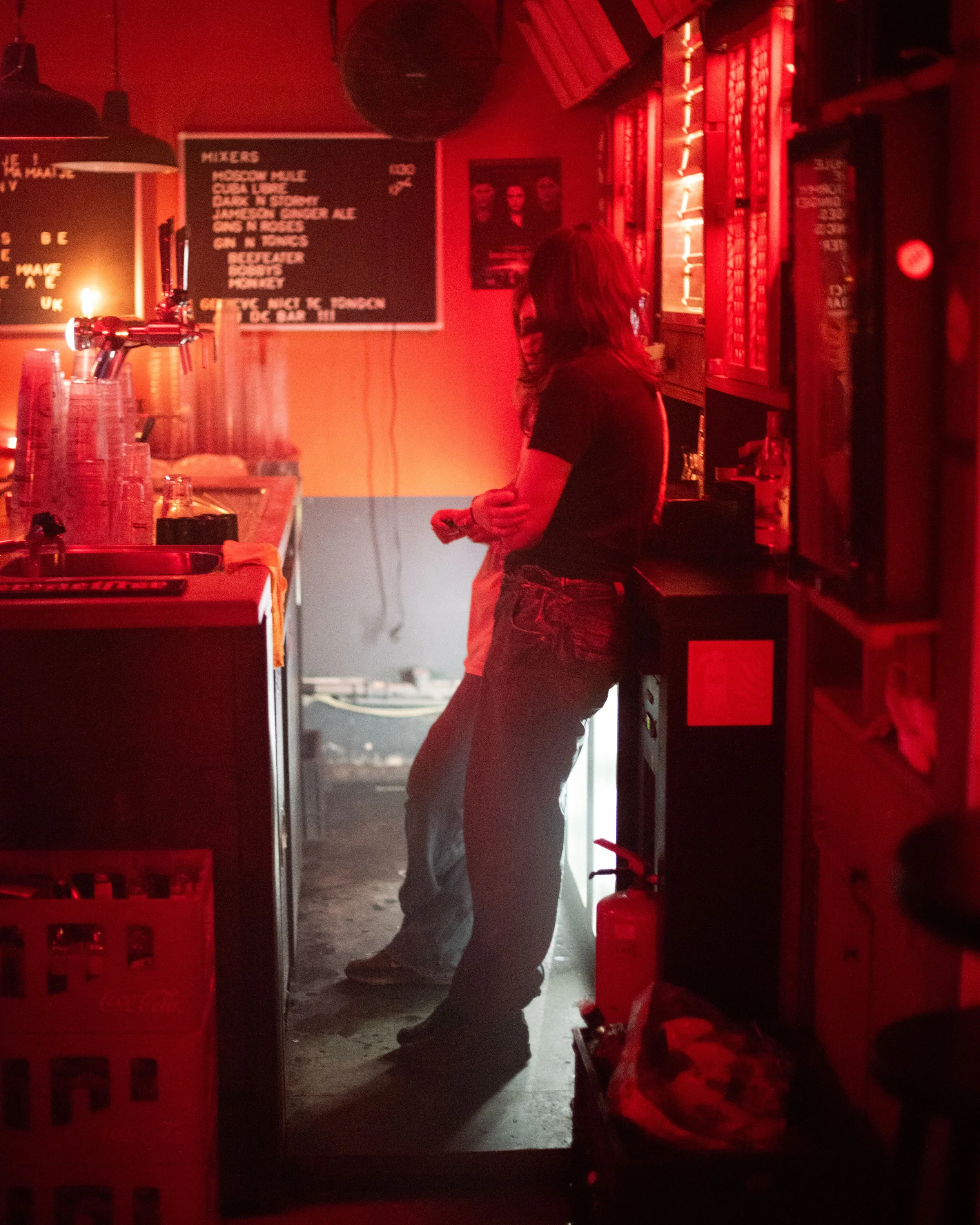 Dimly lit bar with red lighting, person leaning against counter, menu board in background, fire extinguisher on floor, crates stacked nearby.