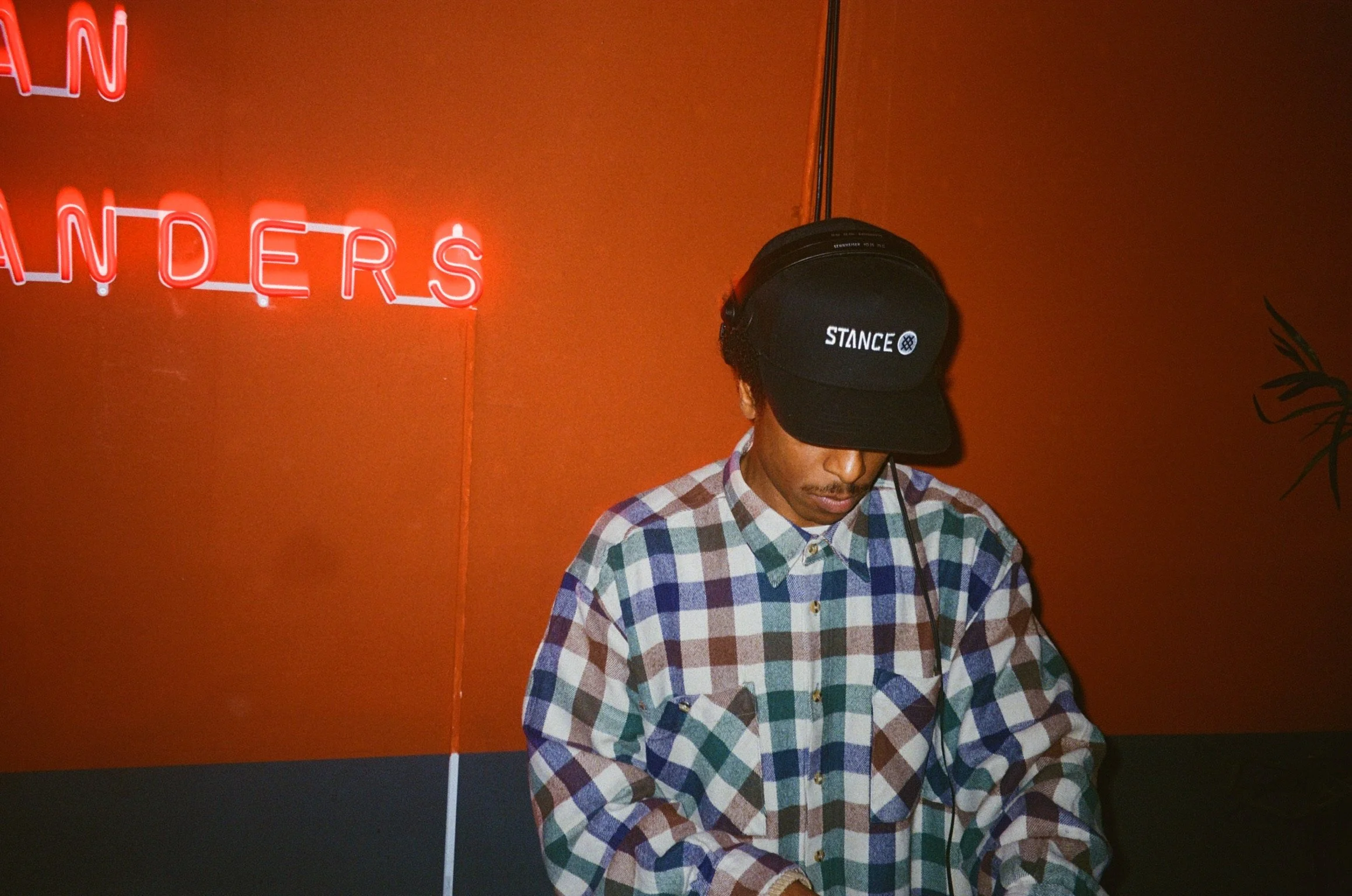 Person wearing a plaid shirt and black cap, standing indoors with red neon letters on an orange wall in the background.