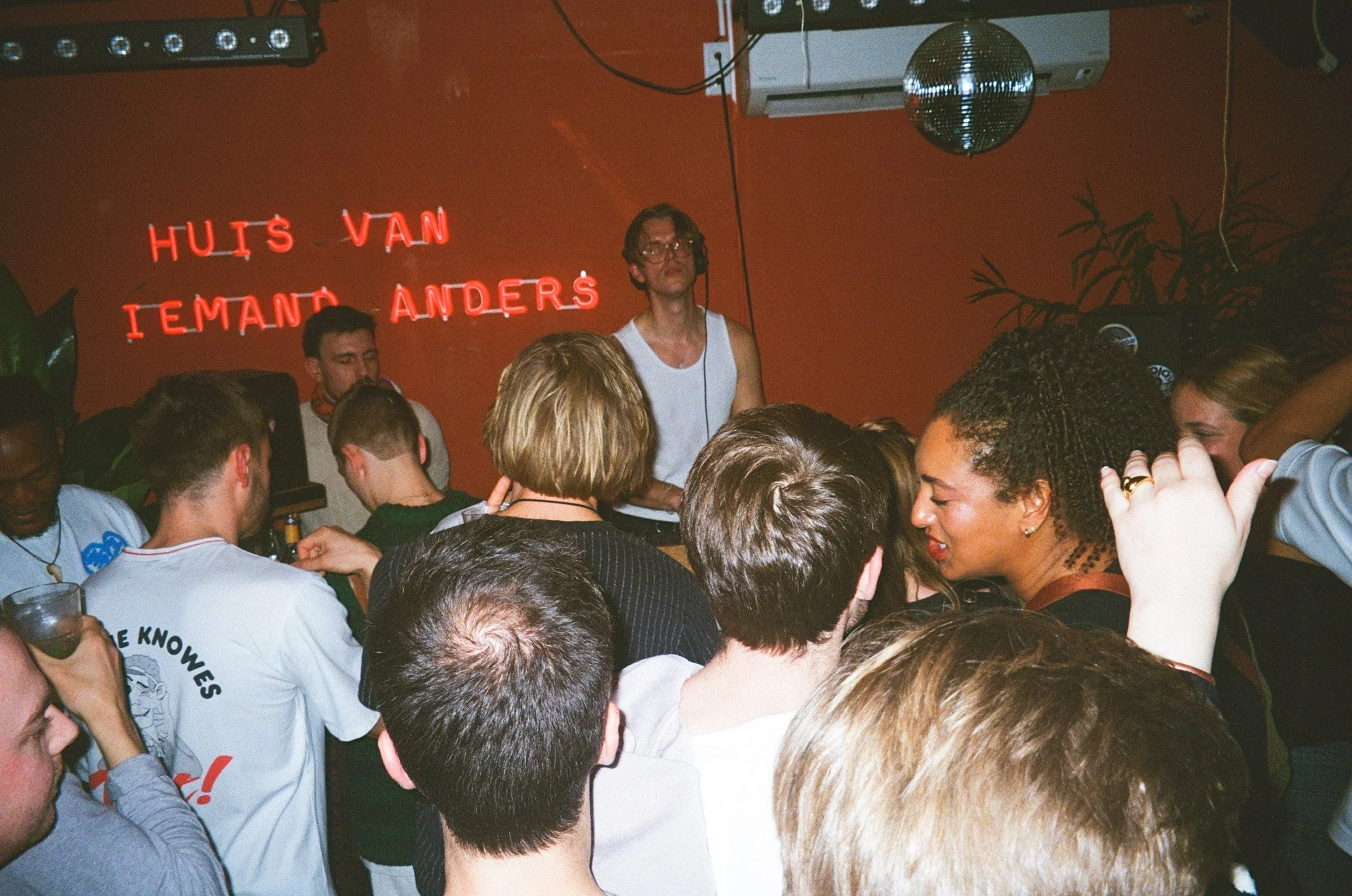 Nightclub crowd with DJ under neon sign "Huis Van Iemand Anders."