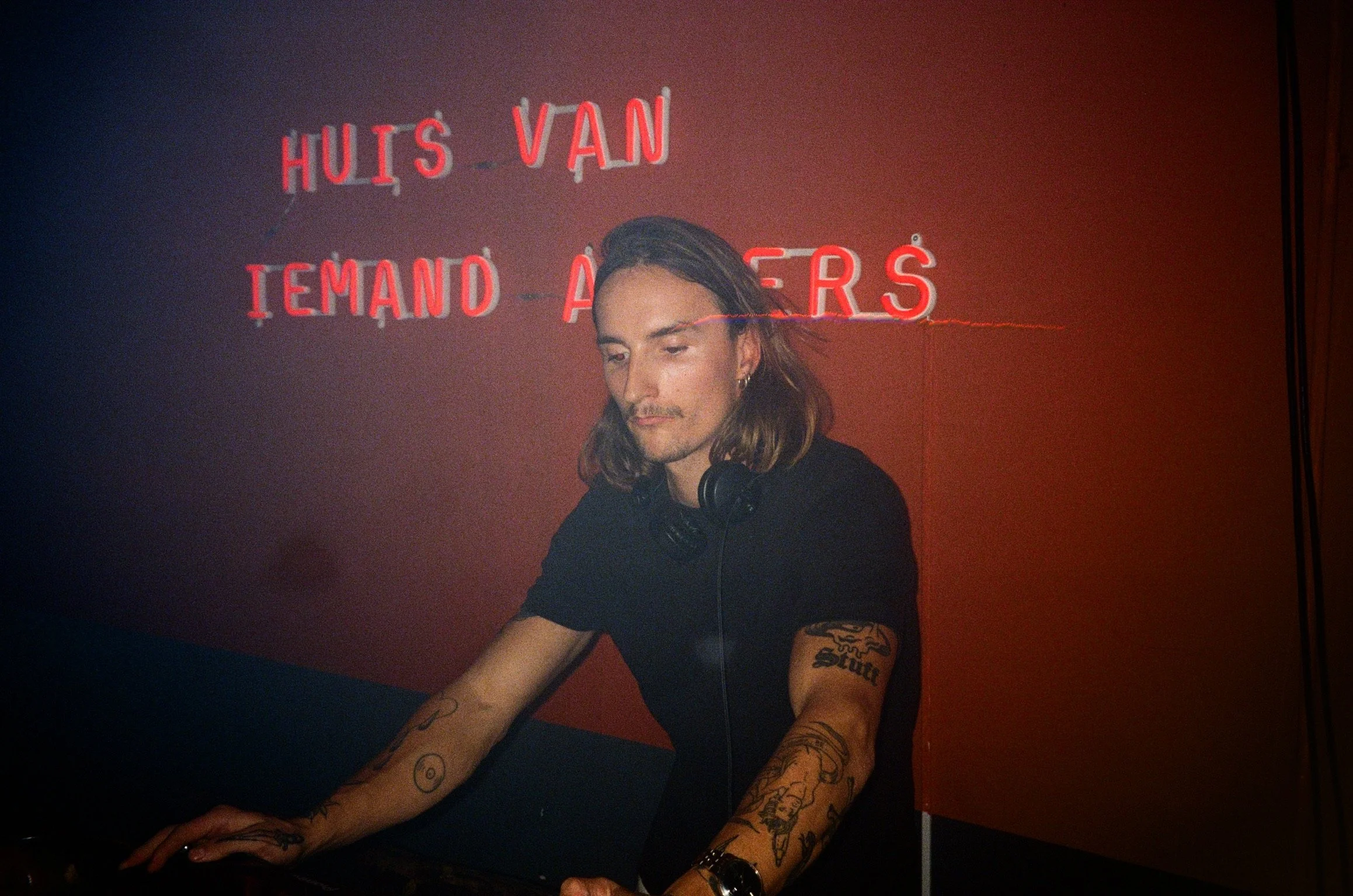DJ performing with headphones, tattoos visible, red neon sign in background.