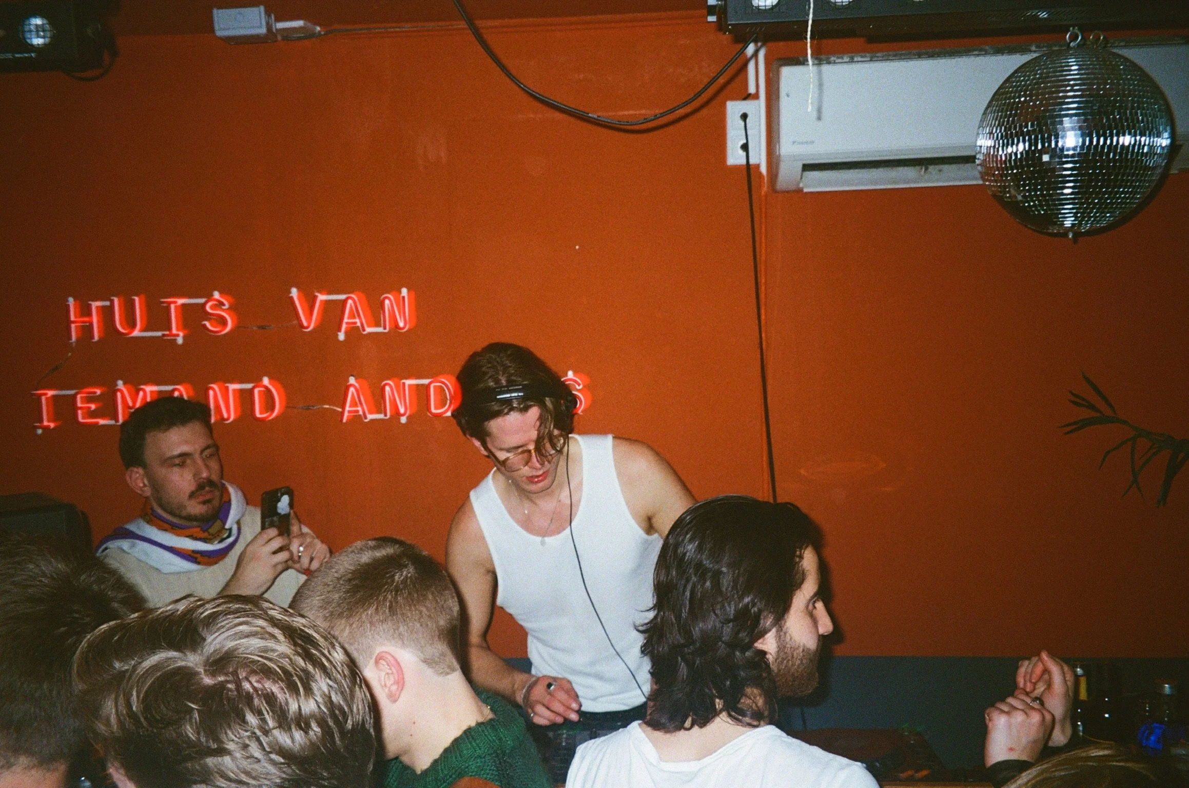 DJ in a club setting with orange walls and neon sign "HUIS VAN IEMAND ANDERS"