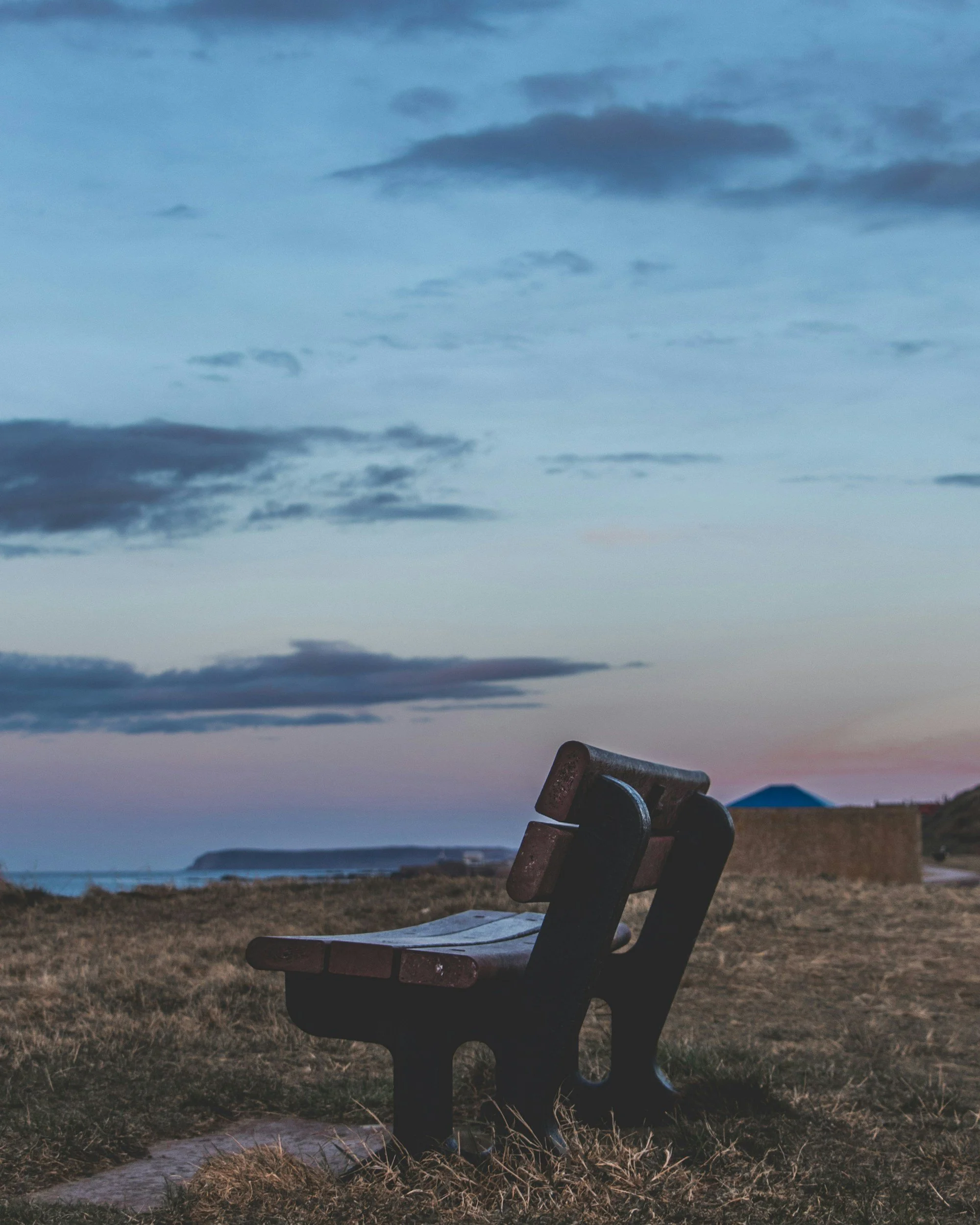 A park bench overlooking a coastal landscape at dusk with a cloudy sky and a distant headland.