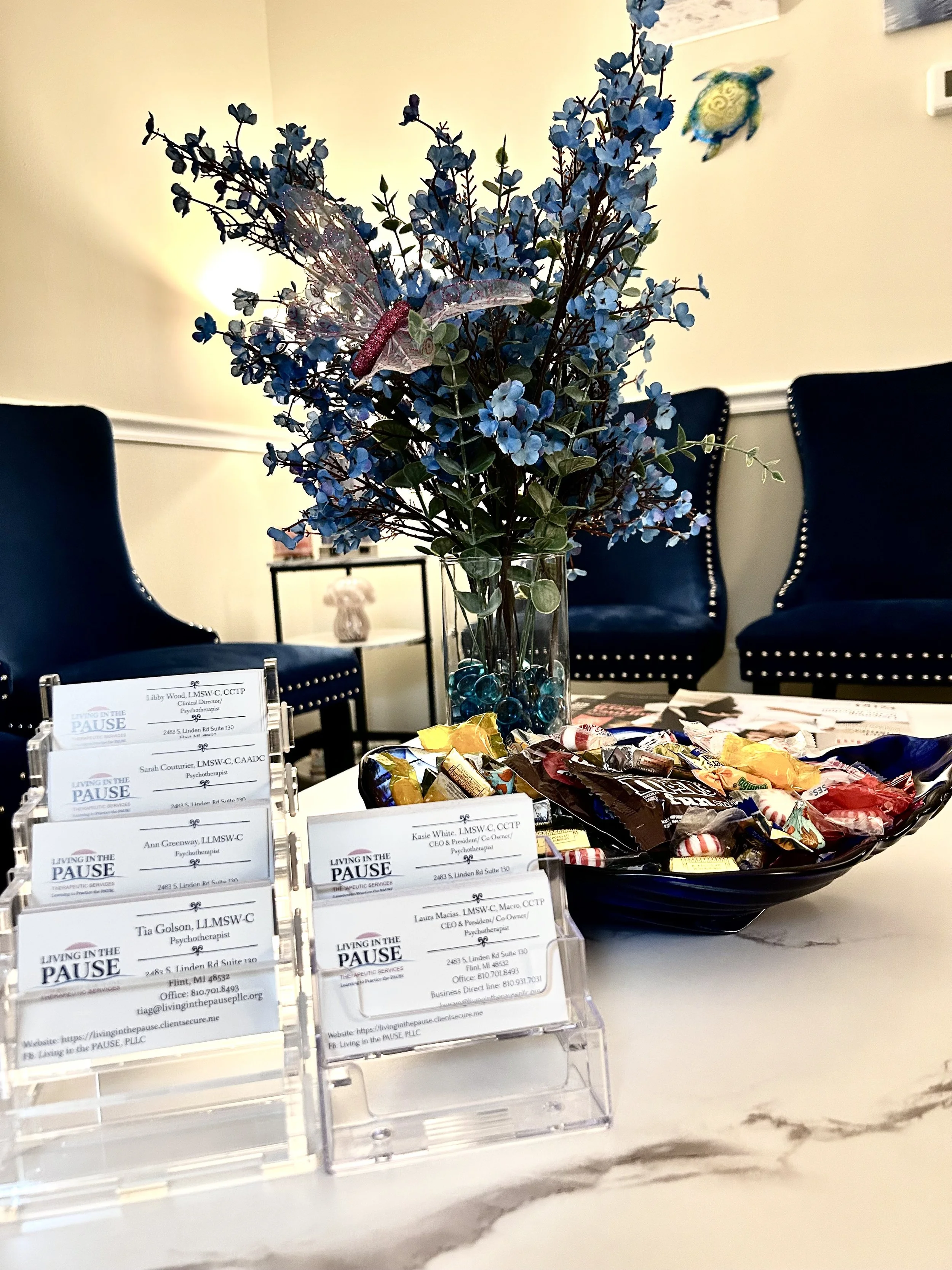 Desk with business cards, a bowl of assorted candies, a vase with blue flowers, a butterfly decoration, and a turtle decoration on the wall.