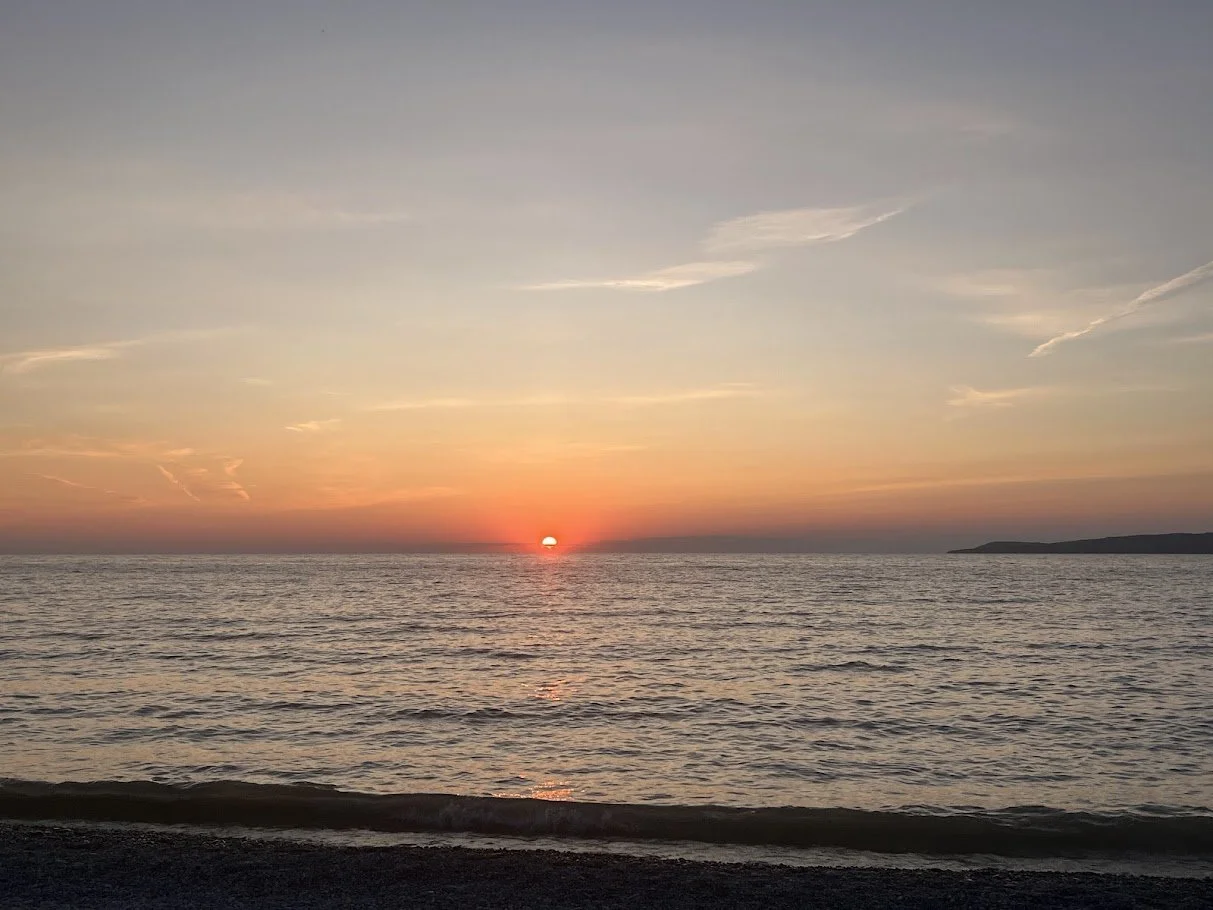 Sunset over the ocean with a calm sea, sparse clouds, and a distant landmass on the horizon.