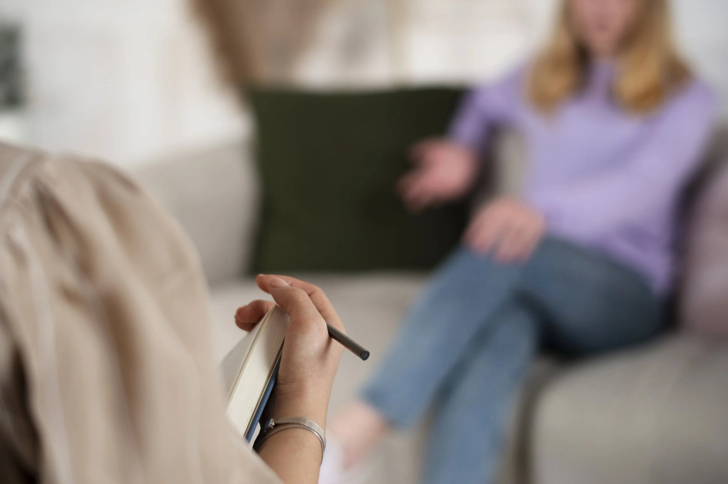A person holding a notebook and pen, with a woman sitting on a couch in the background, engaging in a conversation.