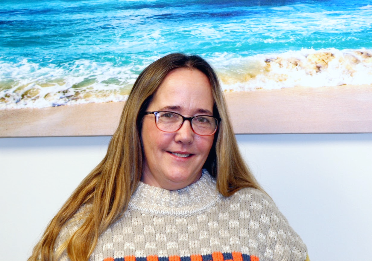 Woman with long brown hair and glasses smiling in front of a beach scene