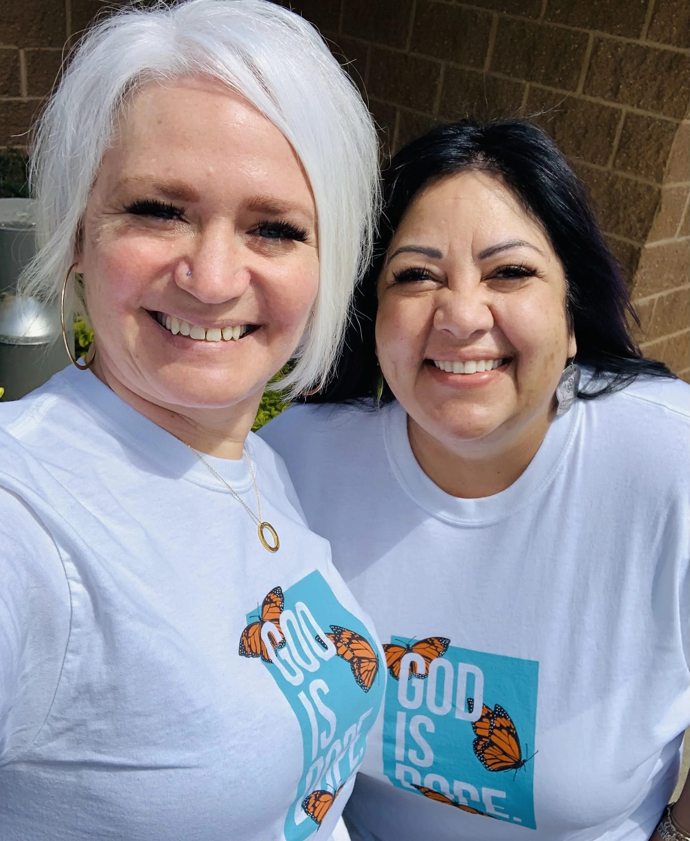 Two women smiling and taking a selfie, wearing white T-shirts with a butterfly and text saying "GOD IS GOOD".