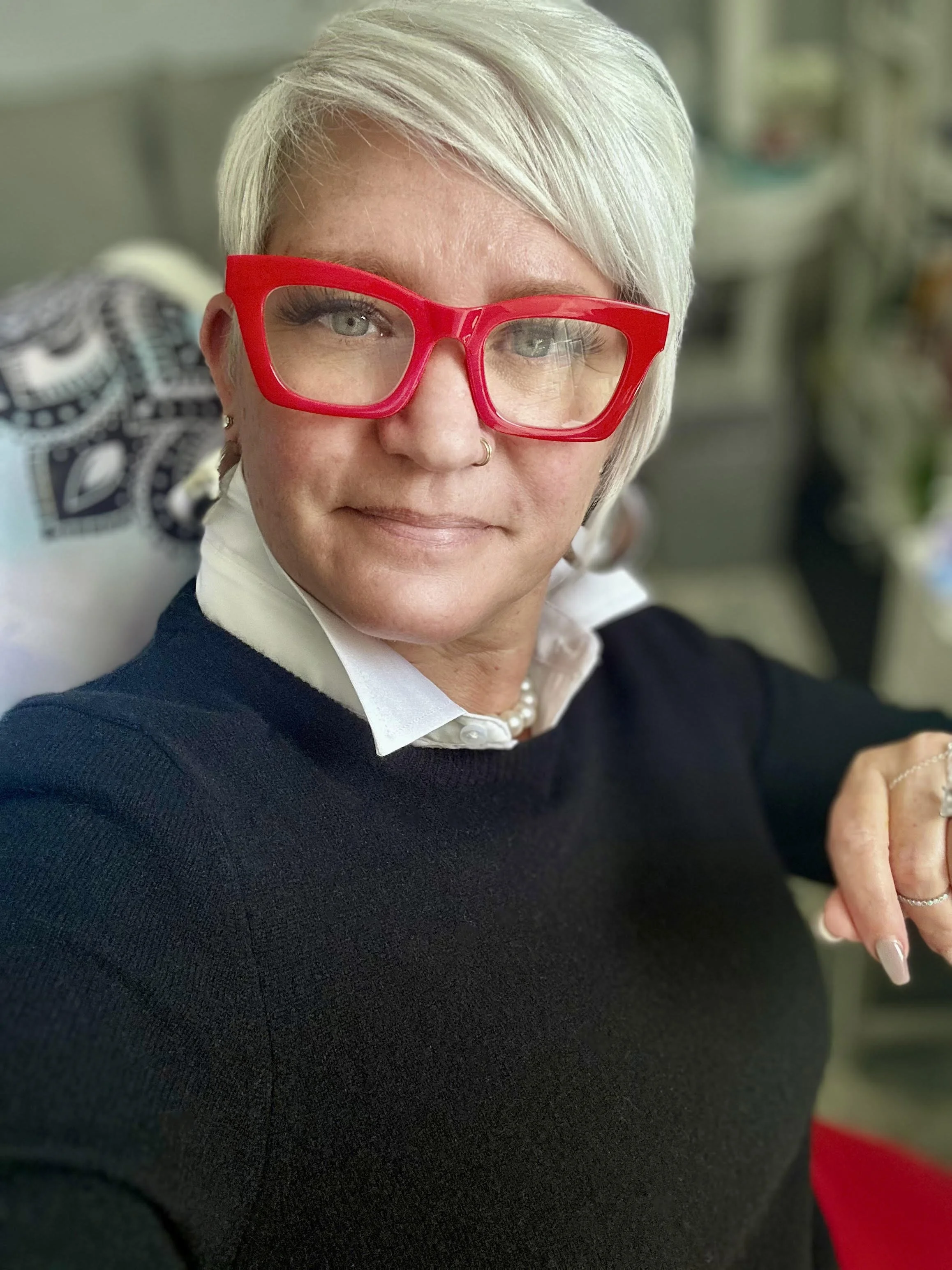 A woman with short, white hair wearing large red glasses, a black sweater, a white collared shirt, a pearl necklace, and earrings, sitting in a room with blurred background.