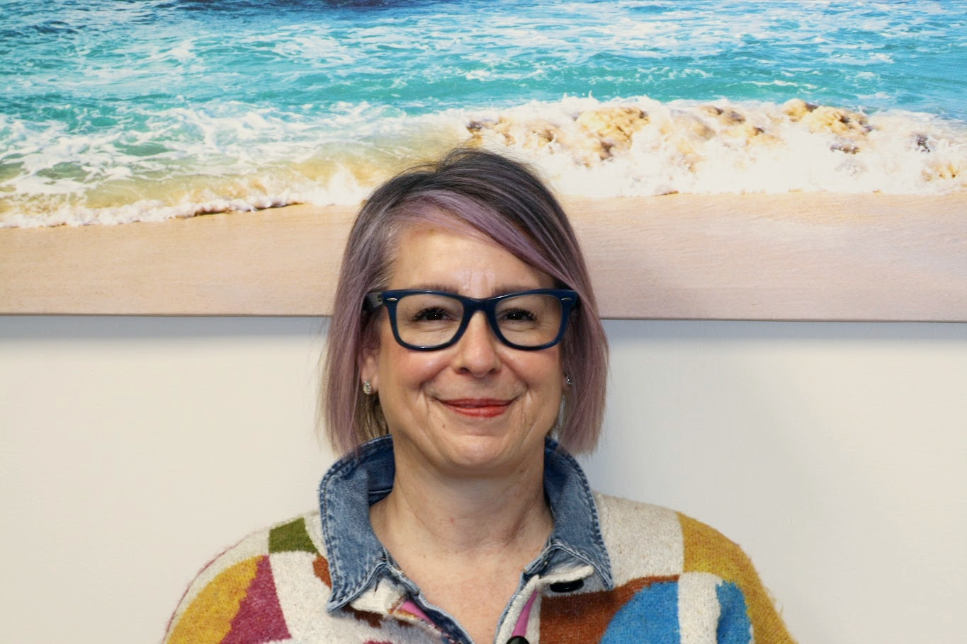 A woman with short purple hair and glasses smiling in front of a beach photograph.