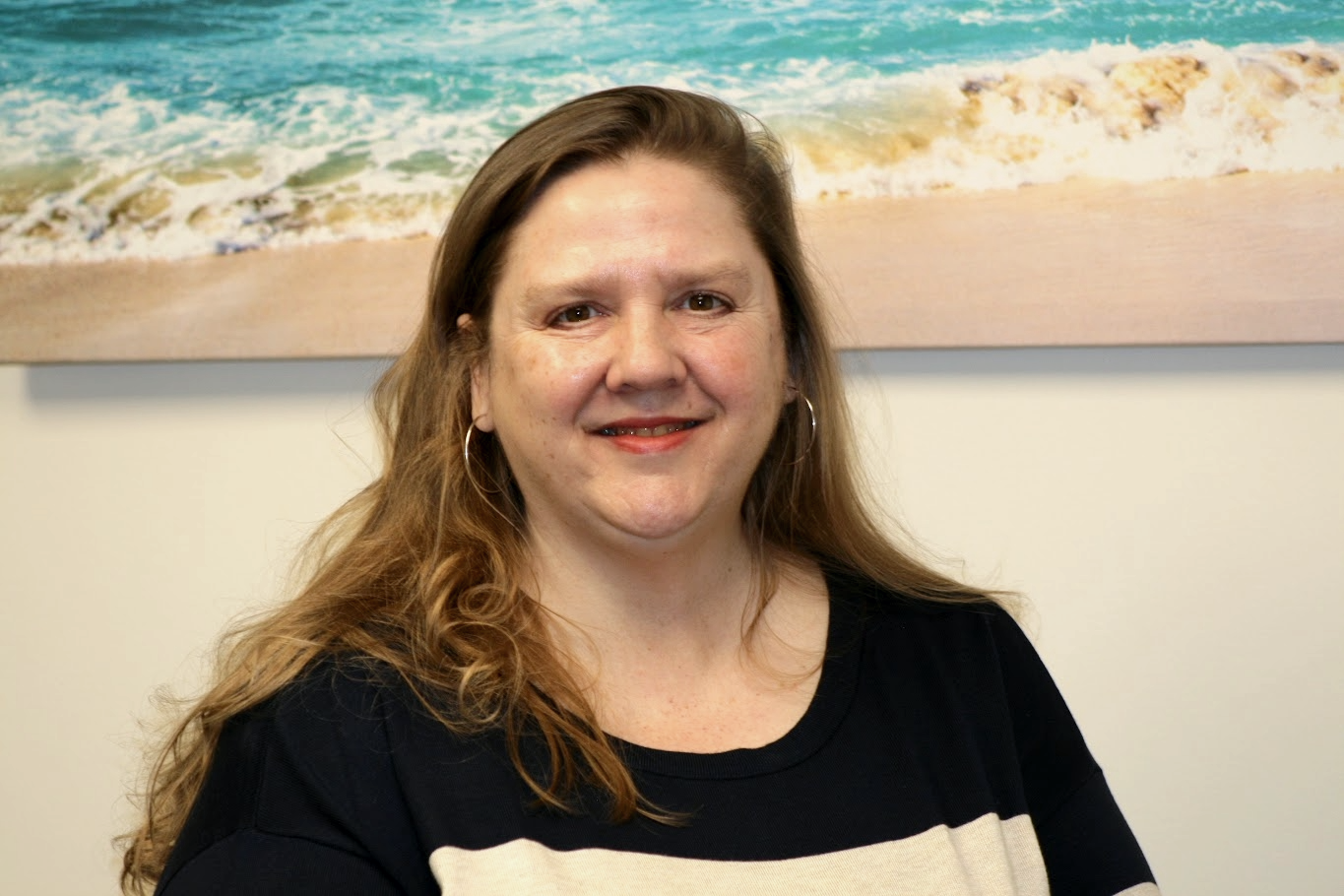 A smiling woman with long brown hair, wearing hoop earrings and a black top, sitting in front of a beach-themed painting.