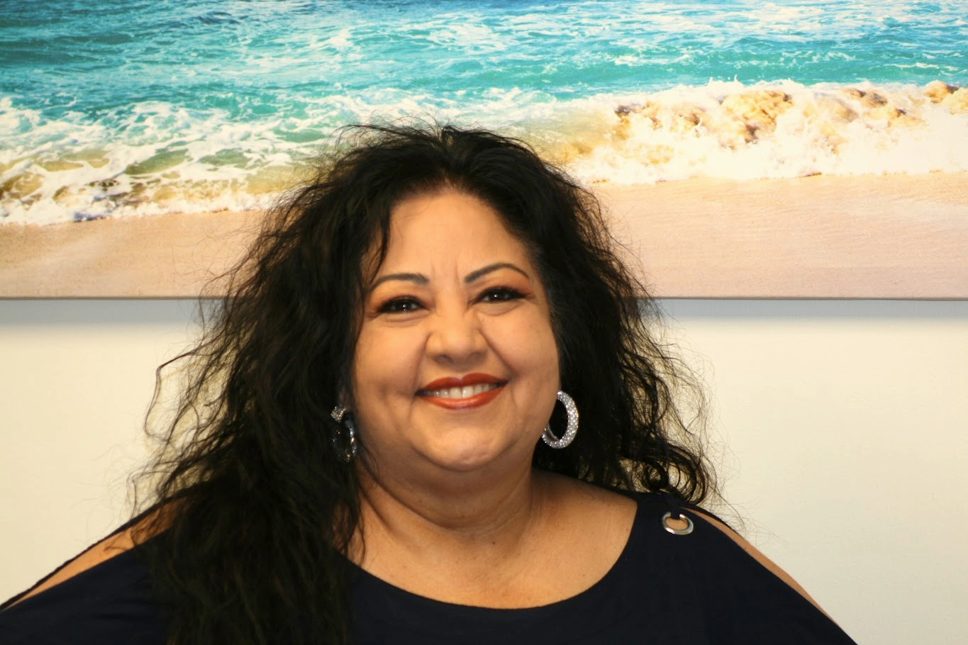 Smiling woman with dark curly hair wearing earrings and a black top, standing in front of a beach scene painting.