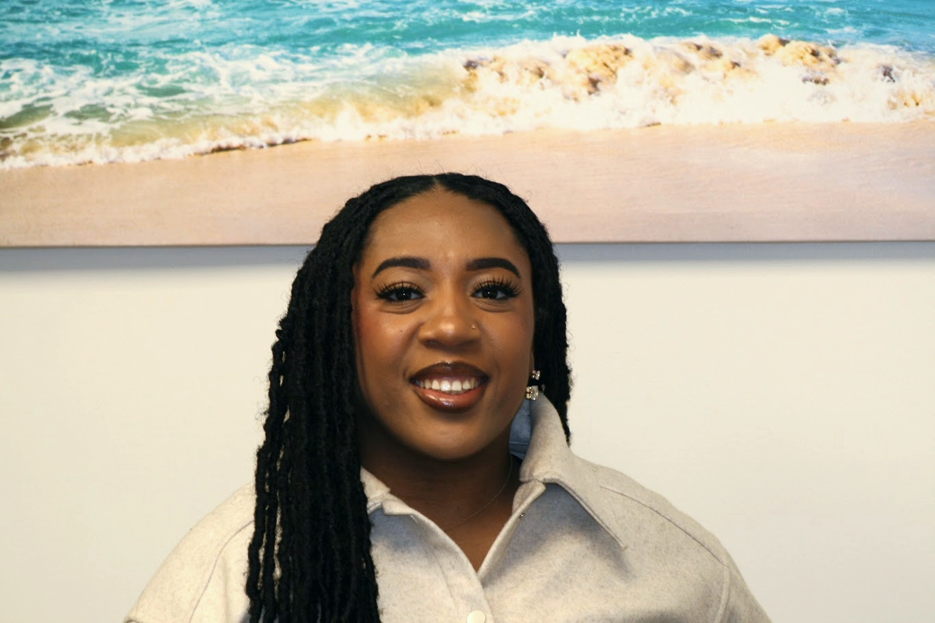 Portrait of a woman with braided hair and earrings, smiling, standing in front of a beach scene painting or photograph.