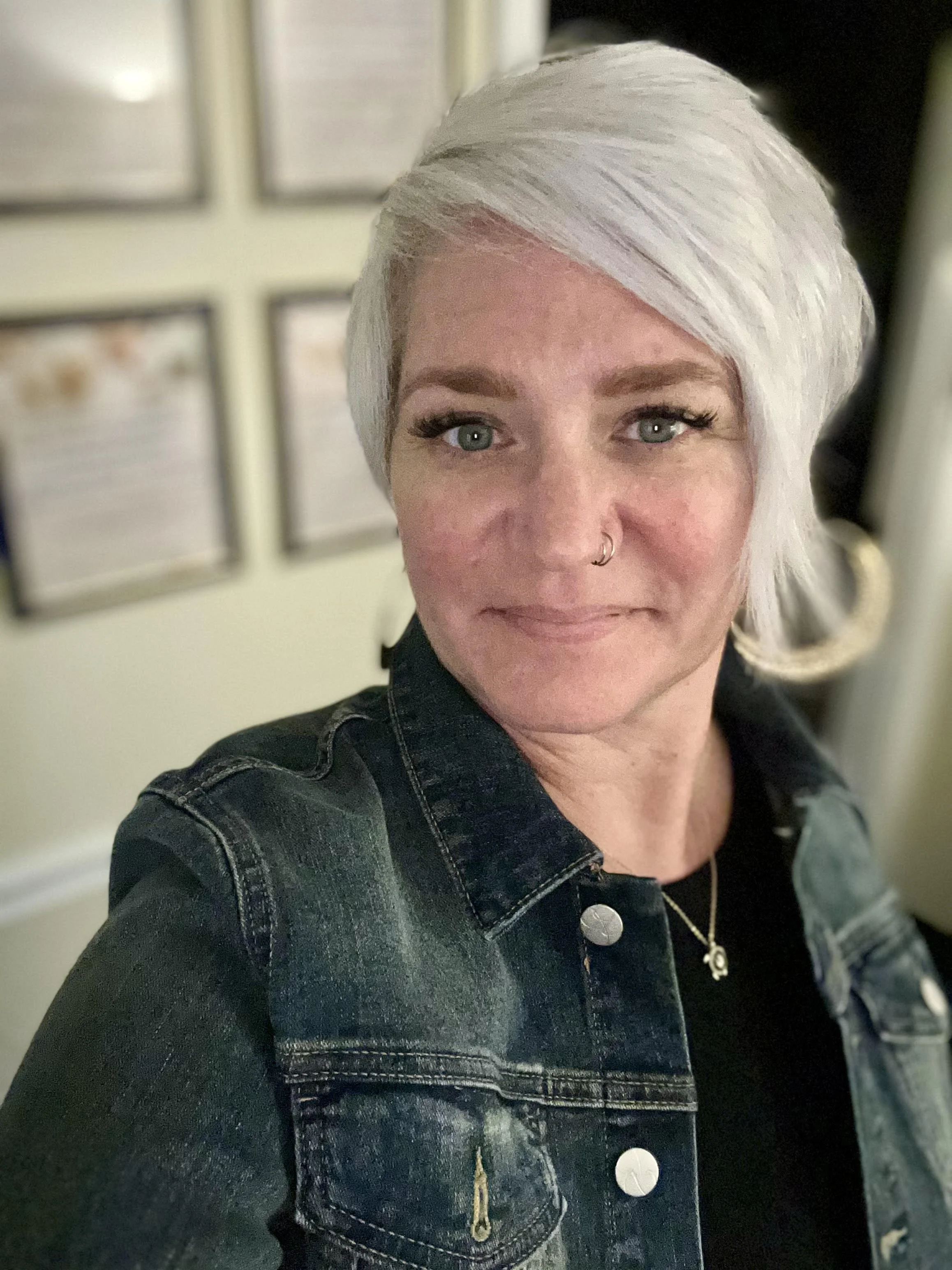A woman with short, platinum blonde hair, wearing a black top and a denim jacket, taking a selfie indoors with framed pictures on the wall in the background.