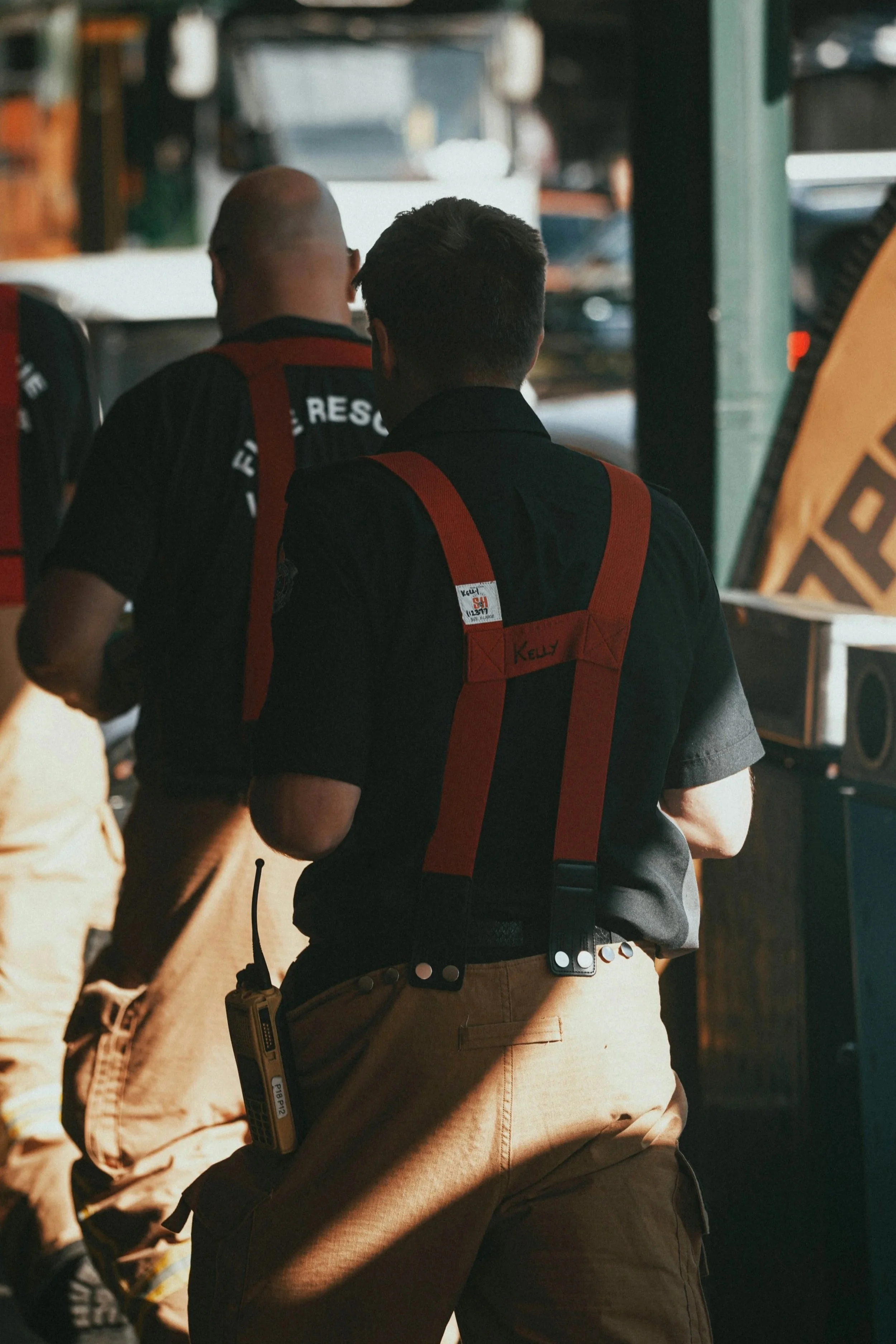 Two rescue workers, one with a label reading 'Kelly,' are seen from behind working at an outdoor emergency scene.