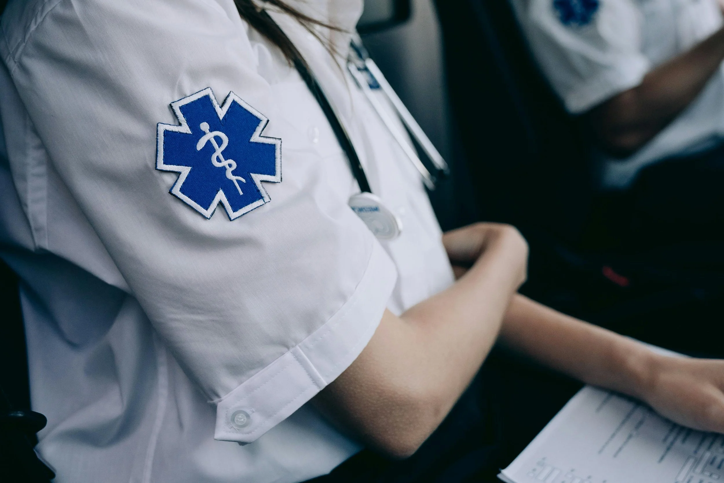 Close-up of a person wearing a white medical uniform with a blue Star of Life patch on the sleeve, a stethoscope around their neck, sitting with arms crossed, holding a clipboard, in a healthcare or emergency response setting.