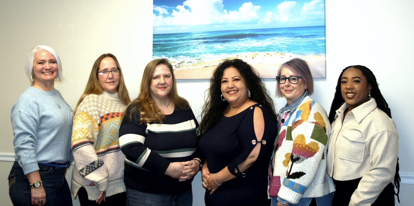 Group of six women standing together indoors with a beach painting on the wall behind them.