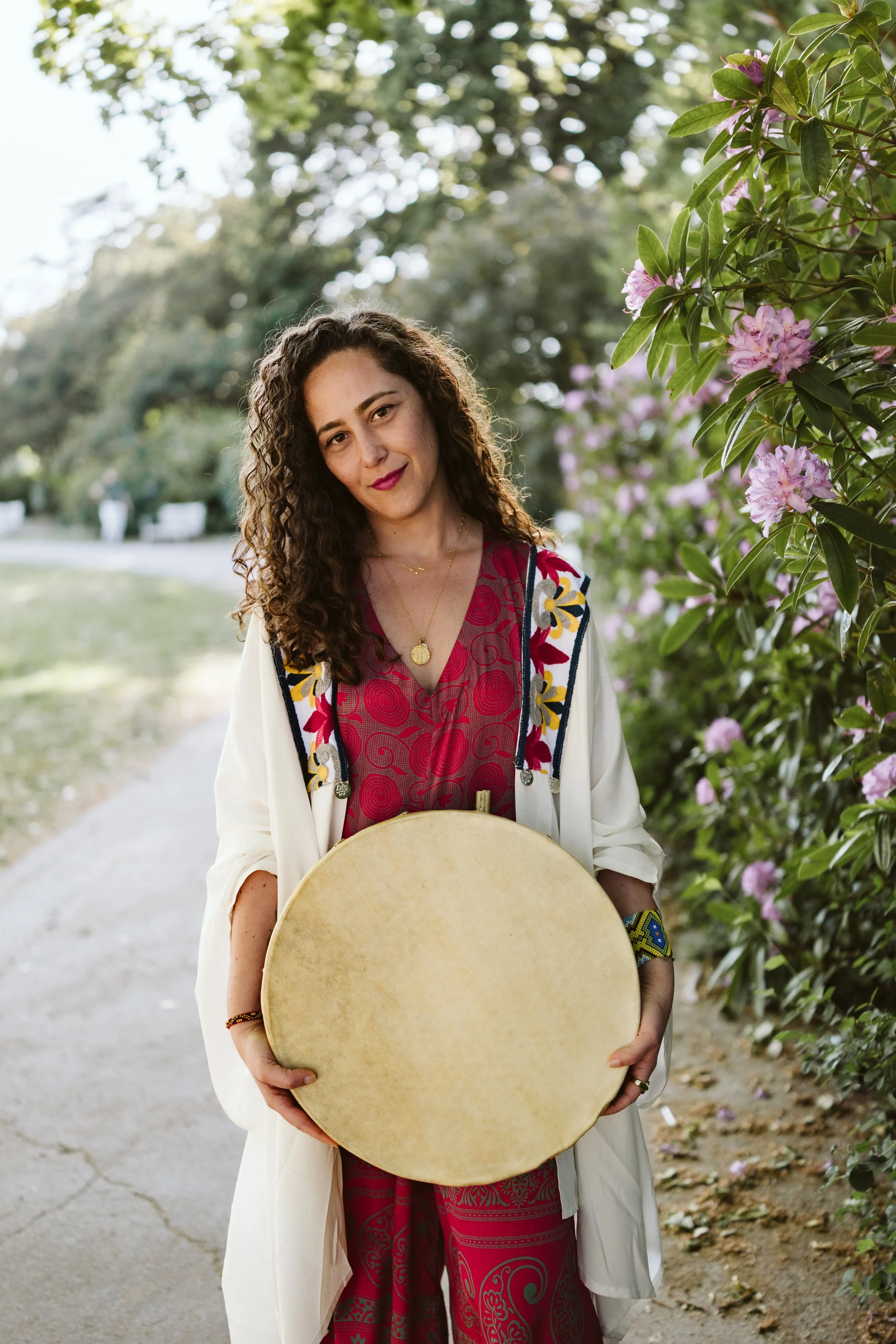 Woman with curly hair, yellow glasses, and feather earrings smiling indoors, with a percussion gong and a candle on a table in the background.