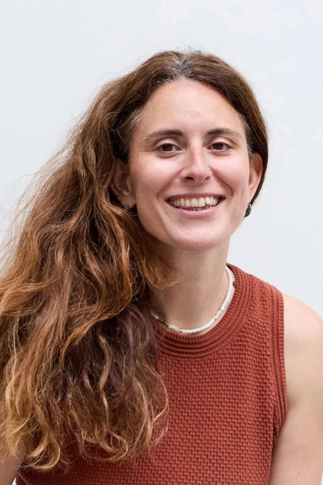 A woman with long, wavy, reddish-brown hair smiling at the camera, wearing a sleeveless red textured top, patterned pants, a pearl necklace, and bracelets, standing against a plain white background.