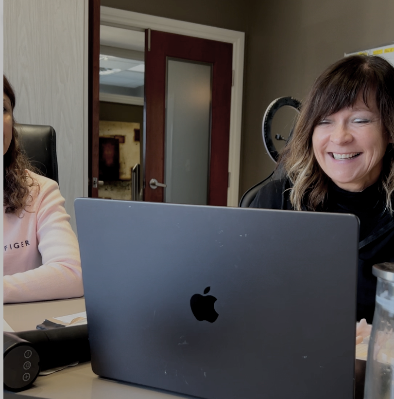 A woman with shoulder-length brown hair and a black top smiling while sitting at a table with a laptop in front of her.